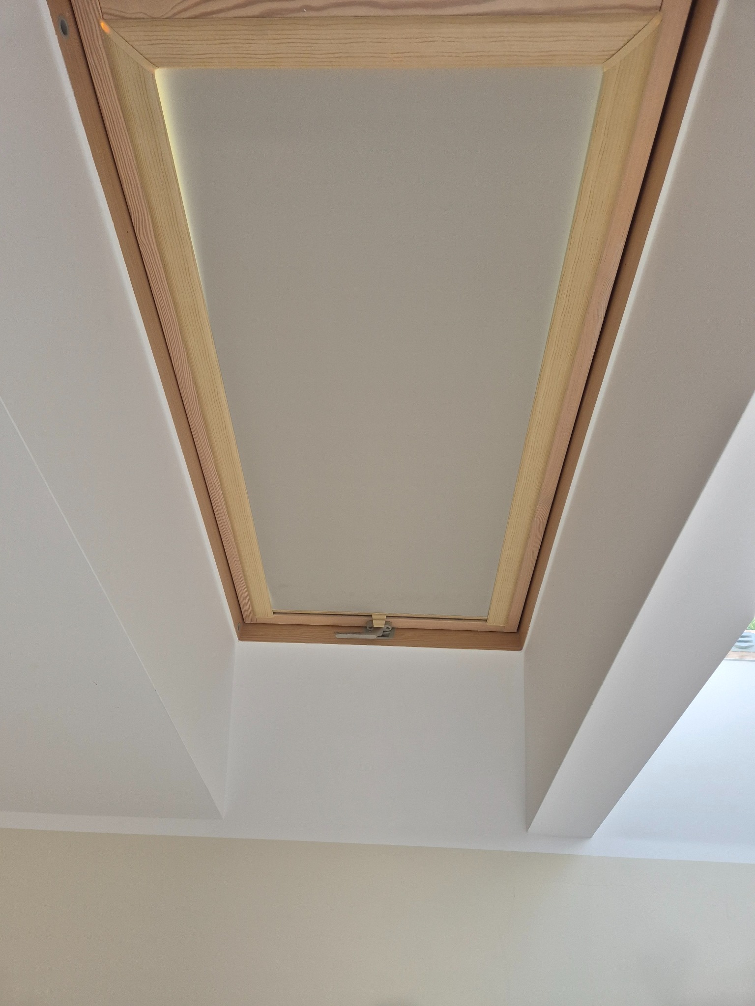 Zamknięte okno dachowe z drewnianą ramą, widoczne od dołu, wkomponowane w biały sufit pod skosem. Widoczny fragment ściany w odcieniu beżu.