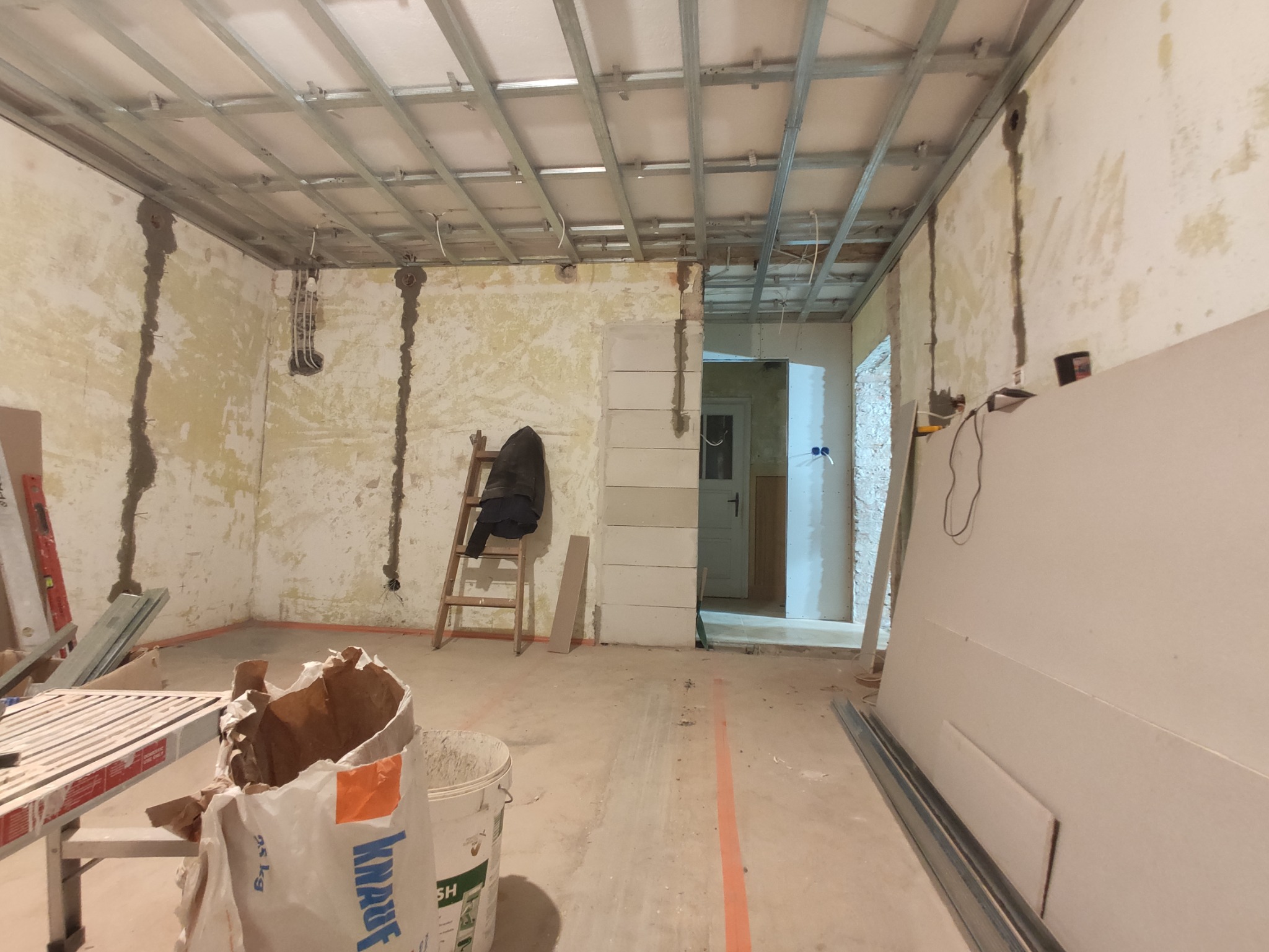 Wnętrze w trakcie remontu: widoczne profile sufitowe, ściany z resztkami starej farby, zaklejone bruzdy instalacyjne, drabina z kurtką, bloczki z betonu komórkowego, płyty gipsowo-kartonowe i worek...