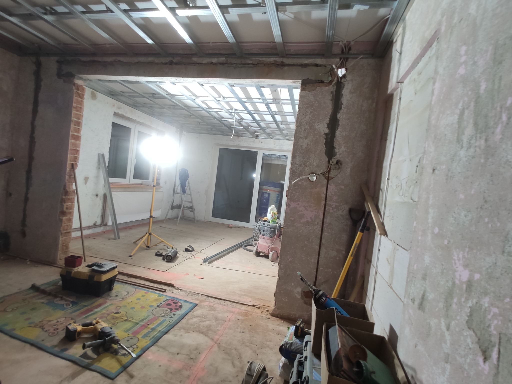 Wnętrze w trakcie remontu: widoczne profile sufitowe, odsłonięte cegły, narzędzia, lampa budowlana na statywie i odkurzacz przemysłowy.