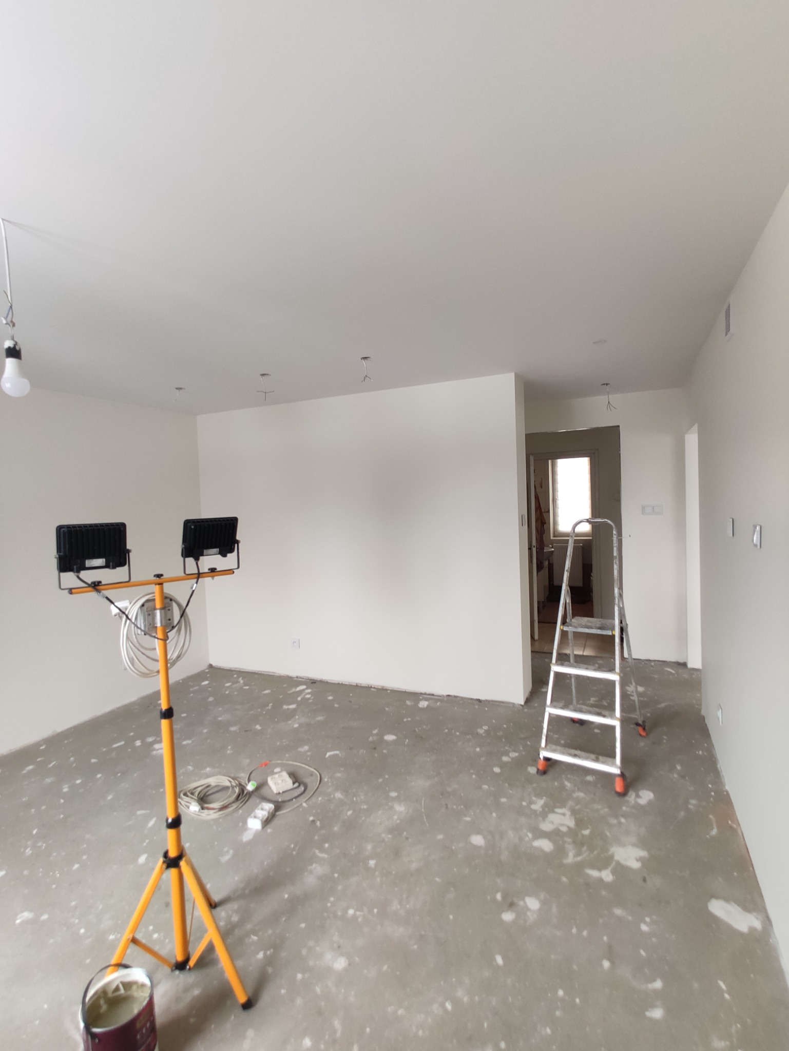 Pokój w trakcie malowania: białe ściany, surowa podłoga z plamami farby, drabina aluminiowa, lampa budowlana na statywie i wiadro z farbą.