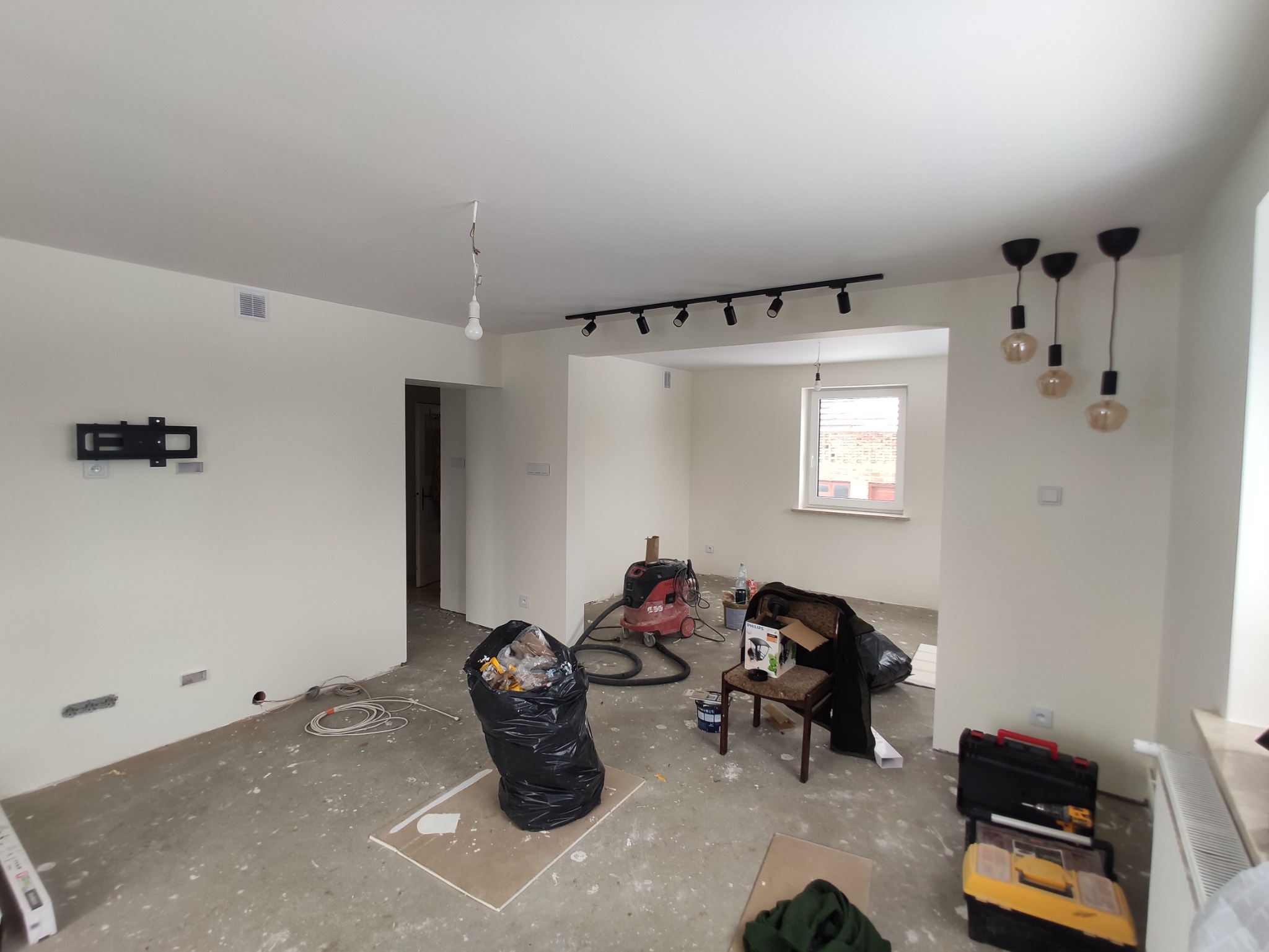 Wnętrze w trakcie remontu: ściany pomalowane na biało, surowa podłoga, widoczne narzędzia, odkurzacz przemysłowy, worki na śmieci i wiszące lampy.