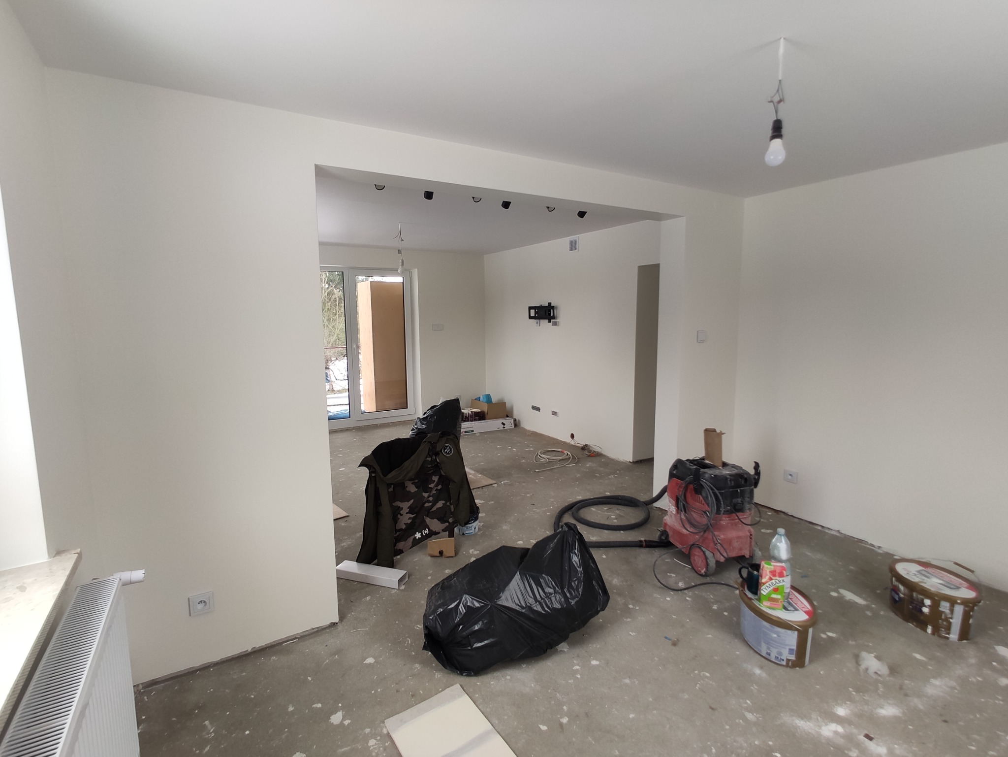 Wnętrze w trakcie remontu: widoczny odkurzacz przemysłowy, worki na śmieci, resztki zaprawy na podłodze, świeżo pomalowane ściany i goła żarówka na kablu.