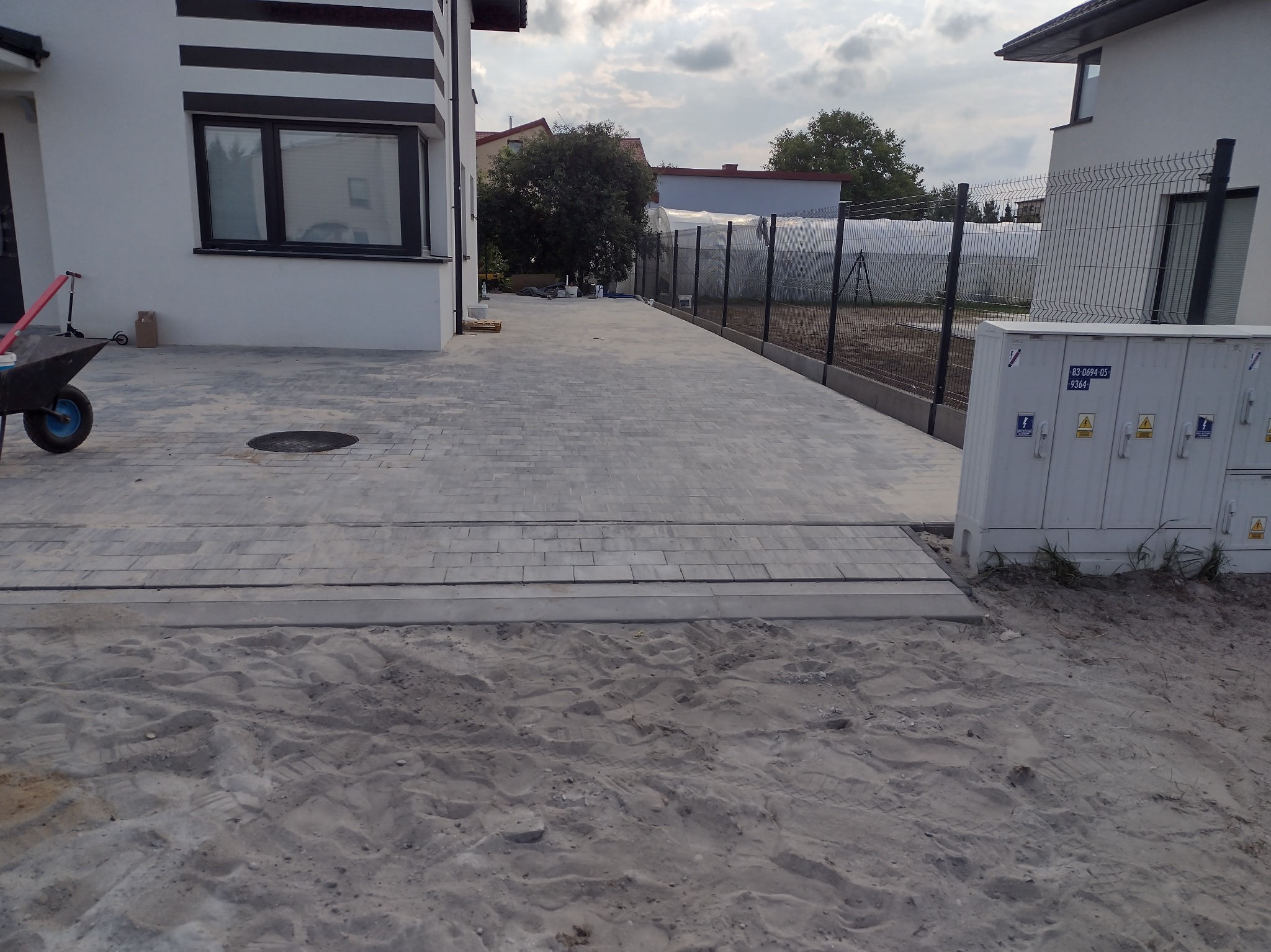 Ułożona szara kostka brukowa na podjeździe przed nowoczesnym domem, widoczna taczka, piasek i skrzynka elektryczna