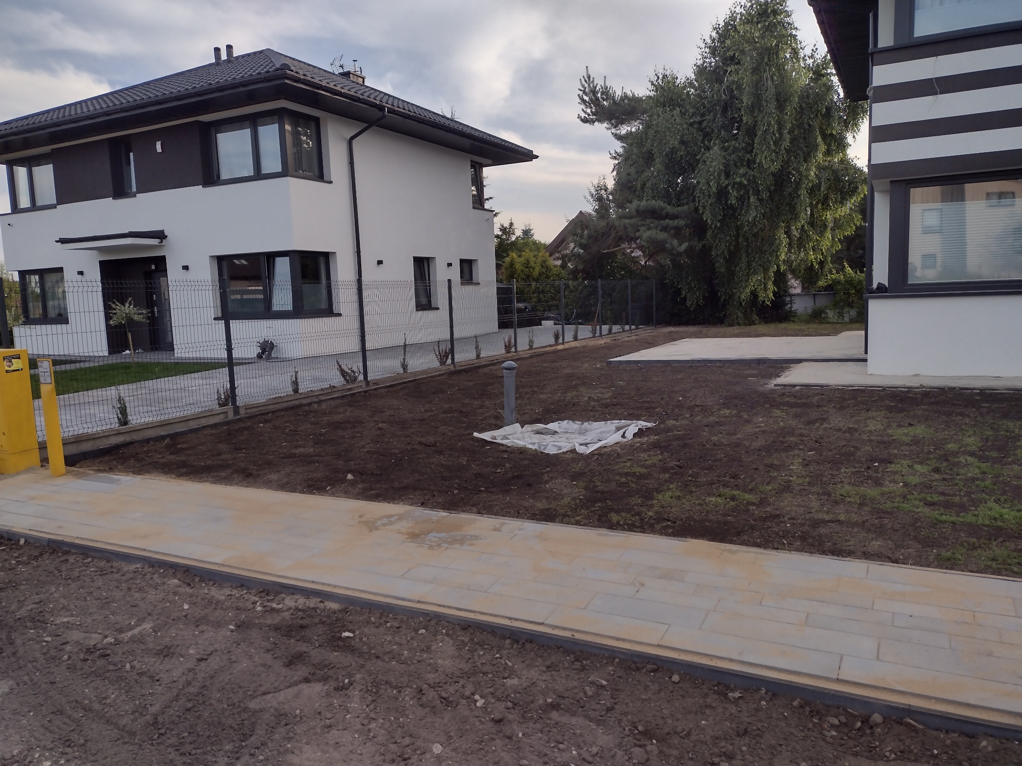 Świeżo ułożony chodnik z kostki brukowej prowadzący do nowoczesnego domu z białymi ścianami i czarnymi akcentami, widoczna przygotowana ziemia pod trawnik i ogrodzenie z siatki.