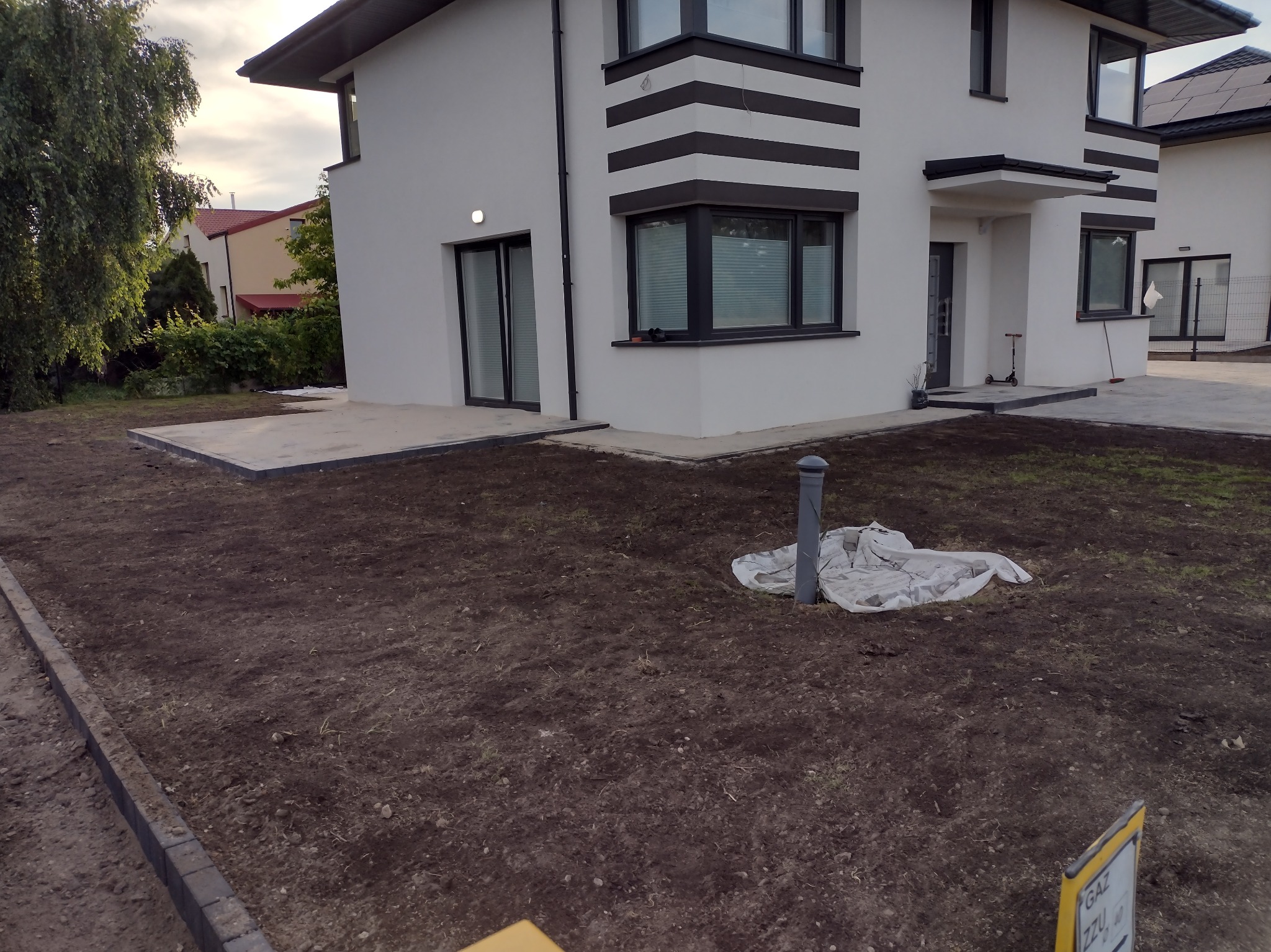 Przygotowany teren pod budowę z widocznymi obrzeżami z kostki, rurą wystającą z ziemi i fragmentem budynku w tle.