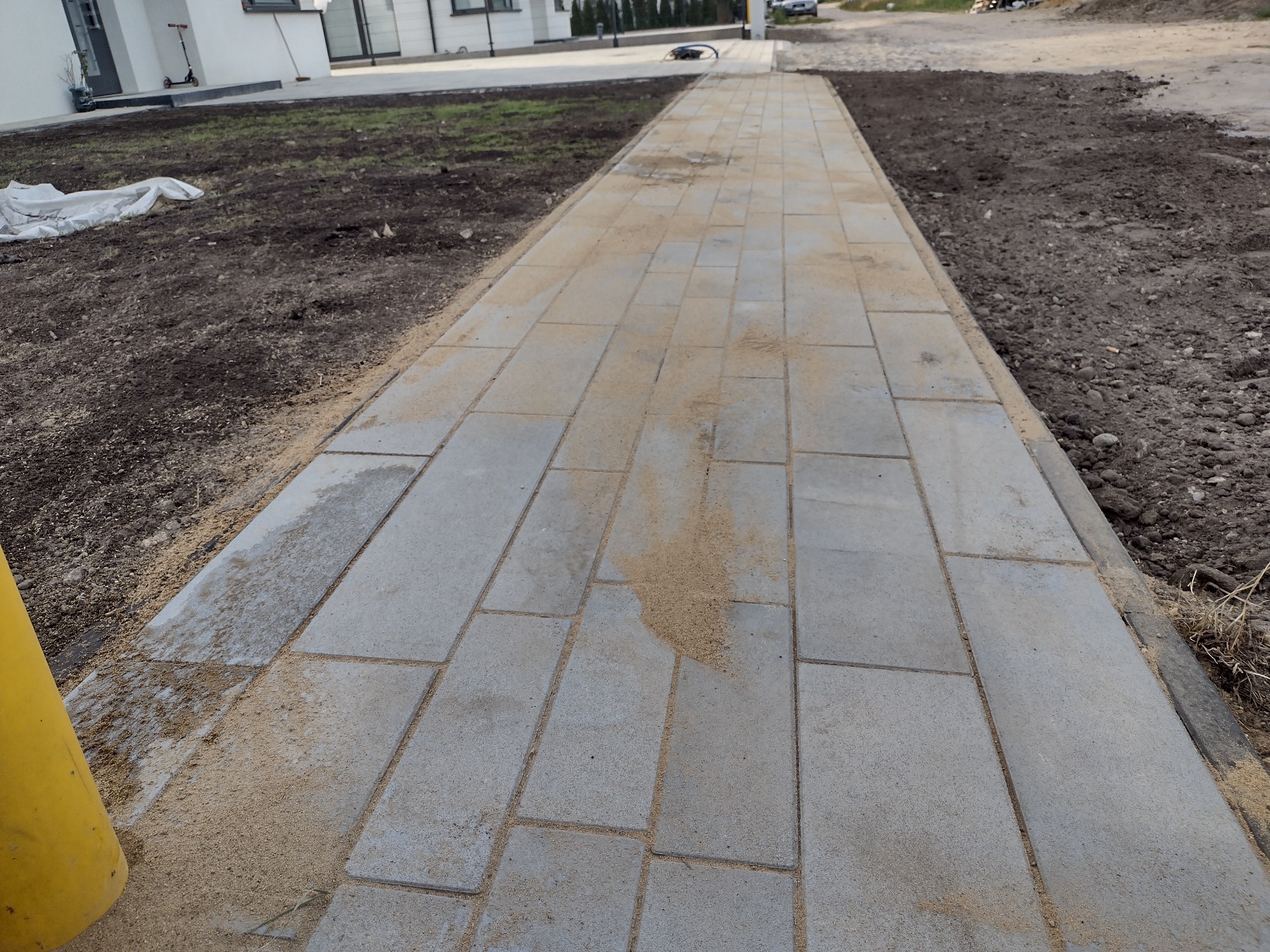 Świeżo ułożona ścieżka z szarych prostokątnych płyt betonowych, fugowana piaskiem, prowadząca wzdłuż nowego domu z przygotowanym pod trawnik terenem.