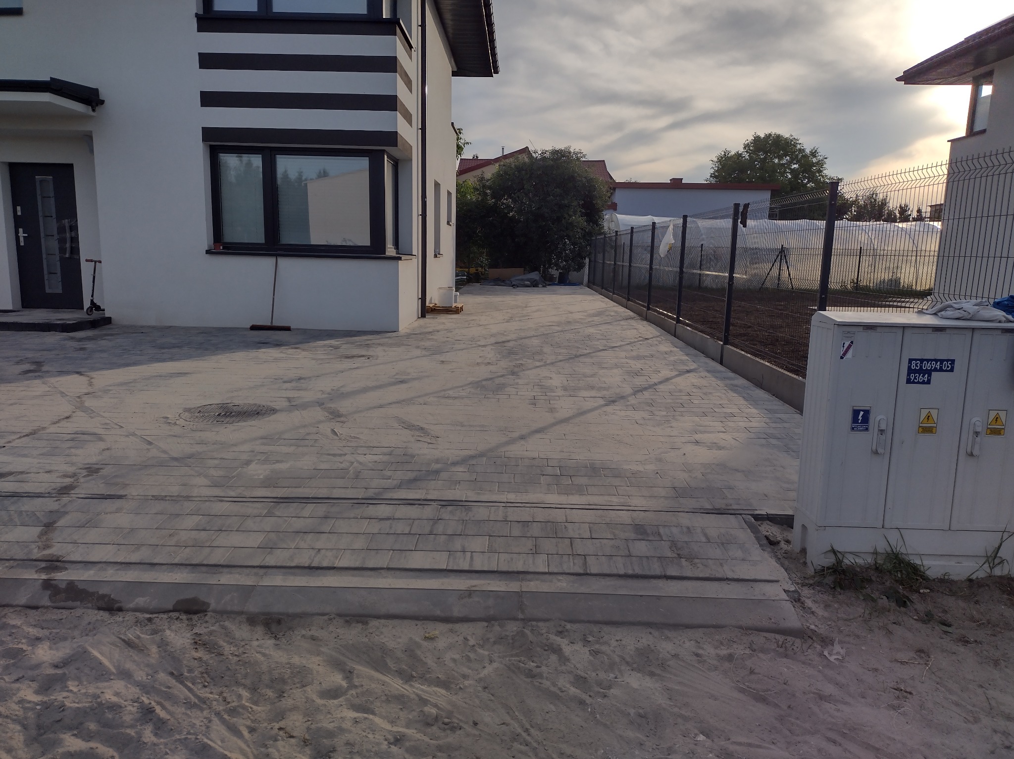 Szara kostka brukowa ułożona na podjeździe do nowoczesnego domu z białymi ścianami i czarnymi akcentami, widoczna skrzynka elektryczna i fragment ogrodu z siatką ogrodzeniową.