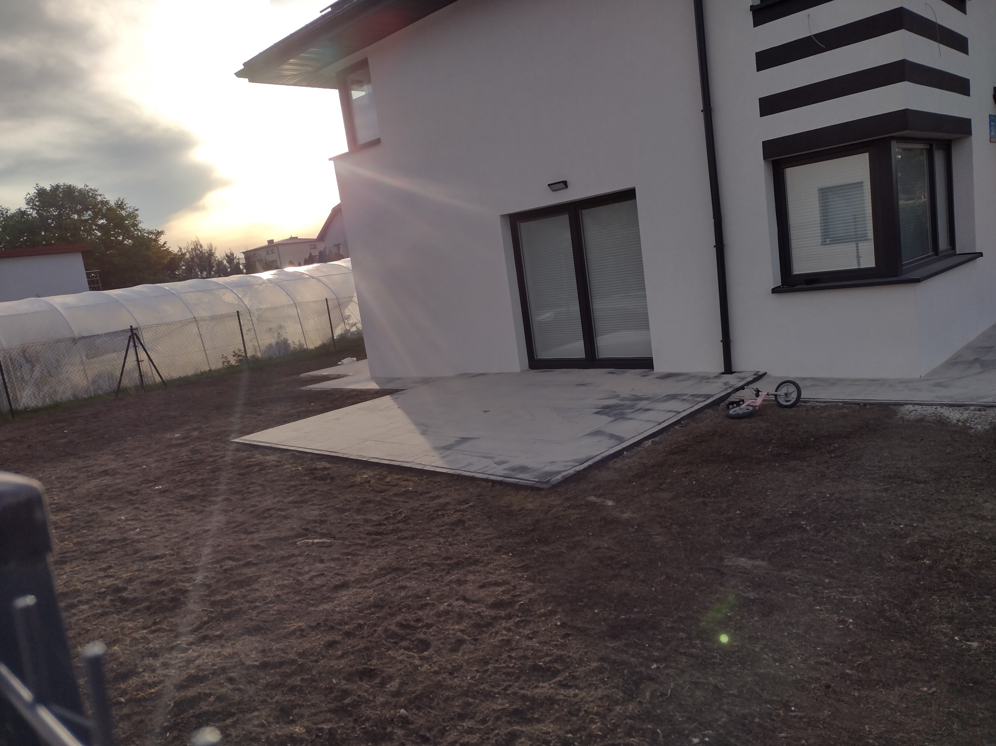 Częściowo ułożona, szara nawierzchnia z kostki przy nowoczesnym, białym domu z czarnymi akcentami i widoczną szklarnią w tle, teren przygotowany pod zasiew trawy.