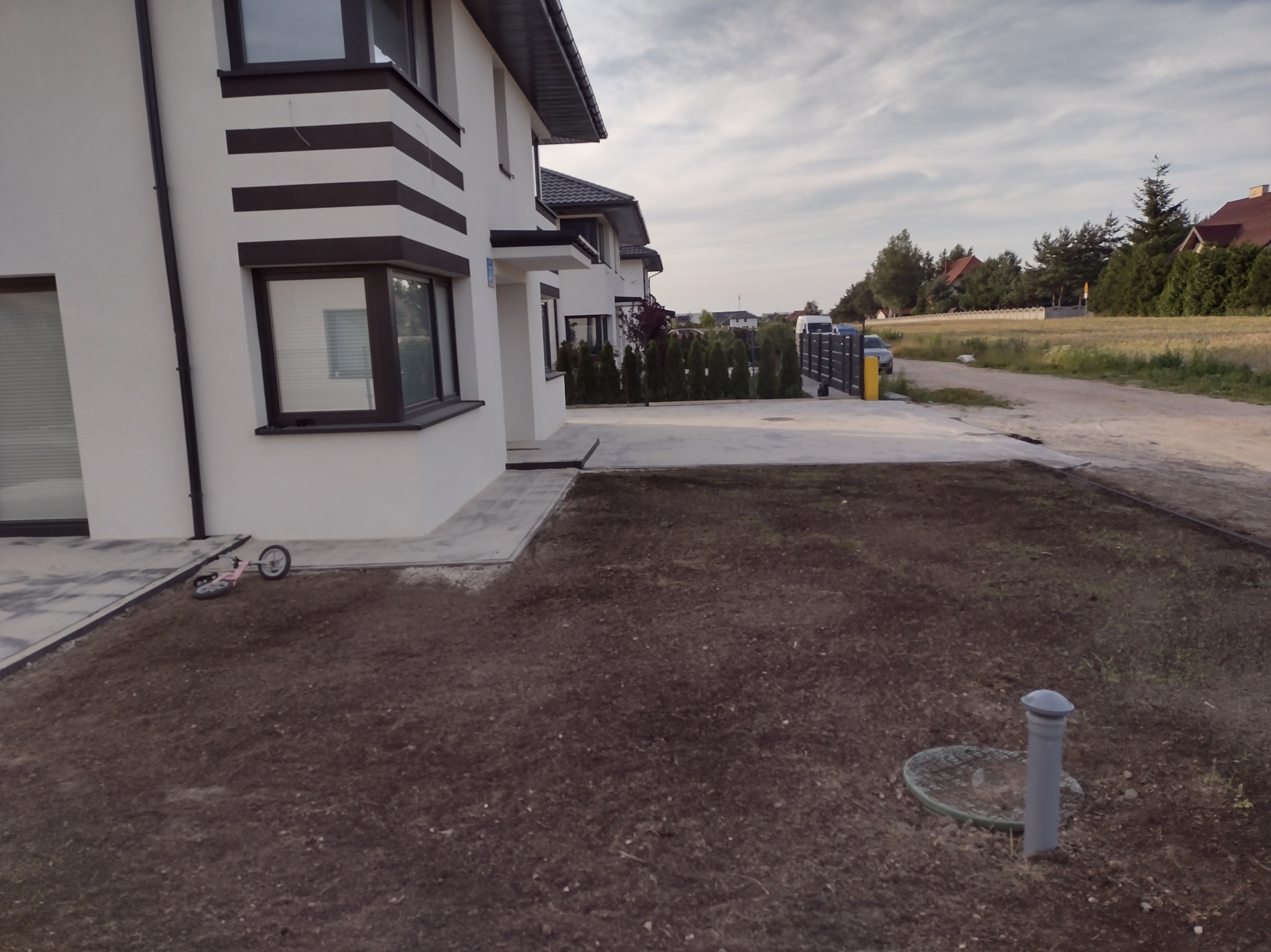 Przygotowany teren pod trawnik przed nowoczesnym domem z szarą kostką brukową przy elewacji, widoczna rura wentylacyjna szamba i dziecięca hulajnoga.
