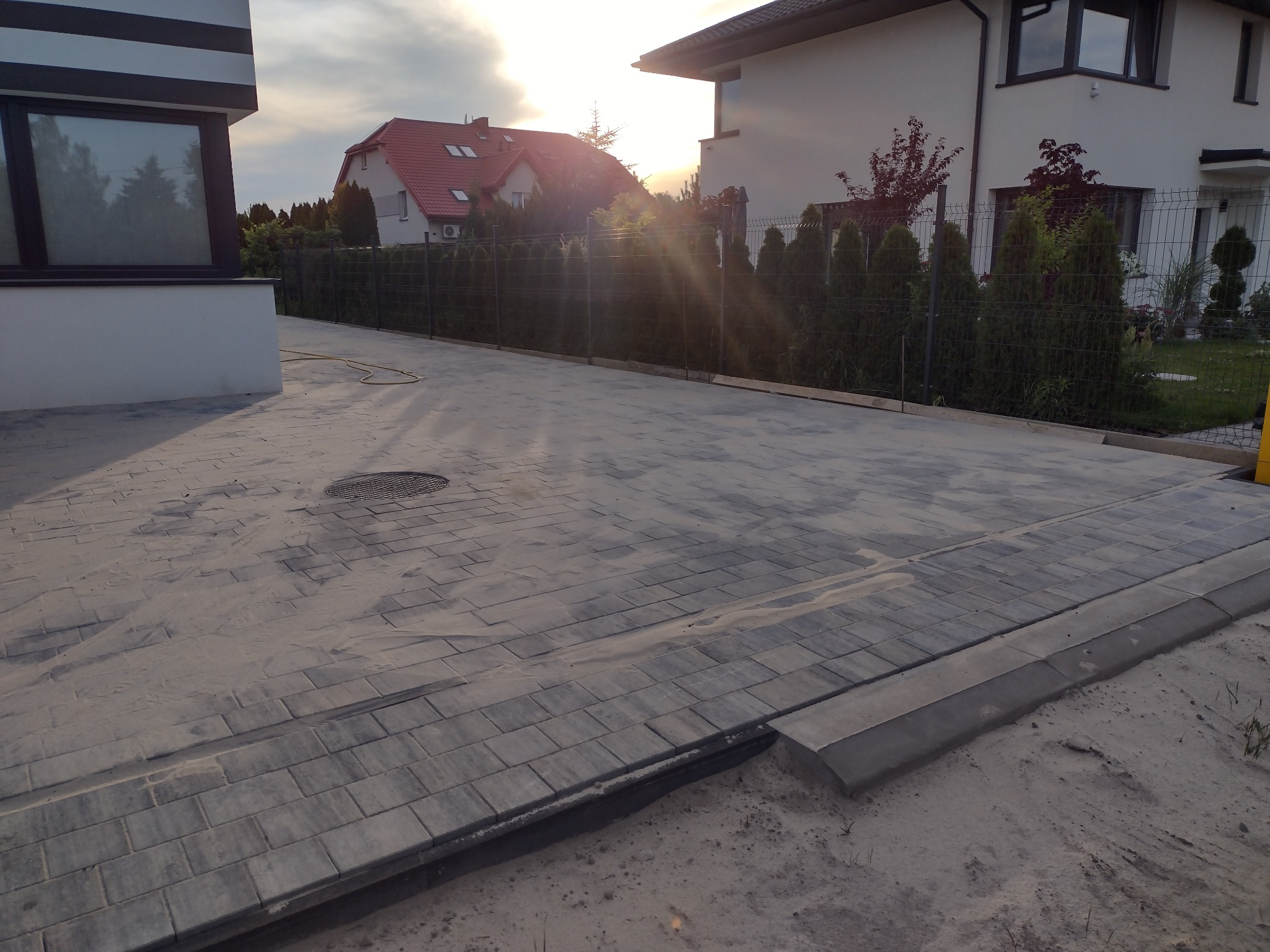 Świeżo ułożona, szara kostka brukowa na podjeździe do nowoczesnego domu jednorodzinnego, z widocznym włazem kanalizacyjnym i piaskiem na powierzchni, w tle ogrodzenie z tujami i zachodzące słońce.