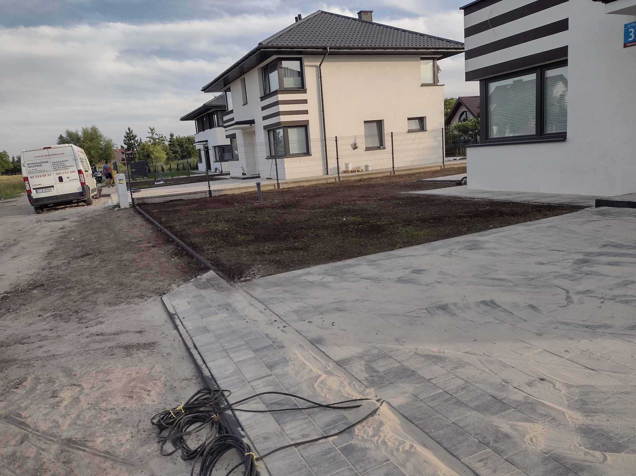 Nowo ułożona, szara kostka brukowa przed nowoczesnym domem z białymi ścianami i ciemnymi akcentami, widoczny kabel elektryczny i przygotowany teren pod trawnik, w tle zaparkowany biały samochód...