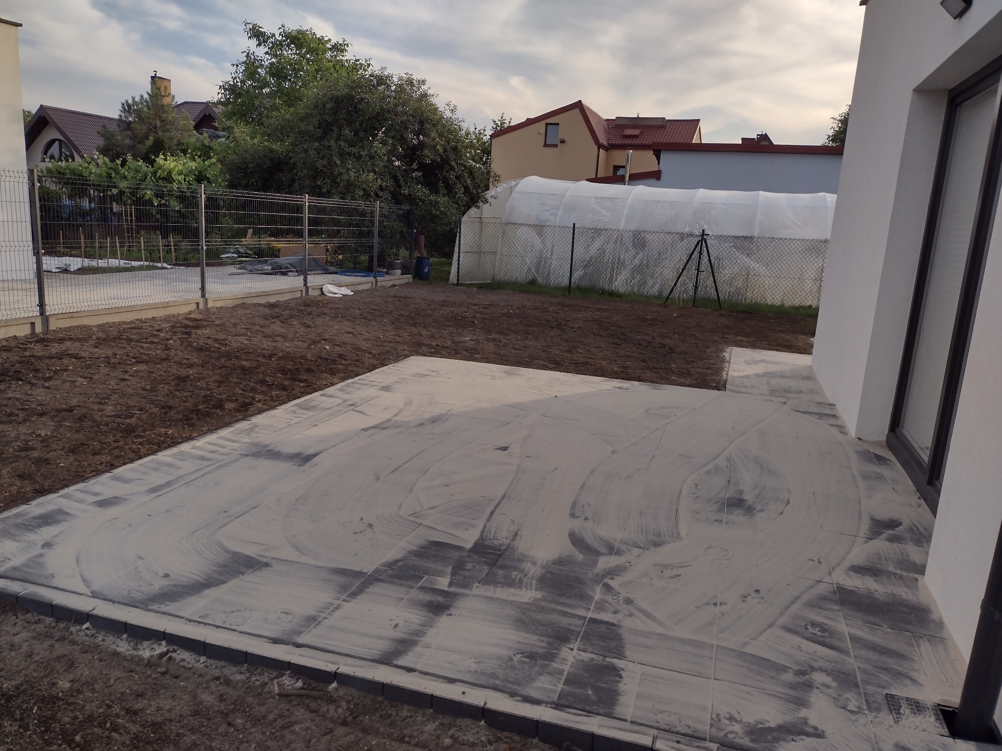 Świeżo ułożona, szara kostka brukowa na przygotowanym podłożu, widoczne ślady po czyszczeniu, w tle ogród z tunelem foliowym i ogrodzeniem.