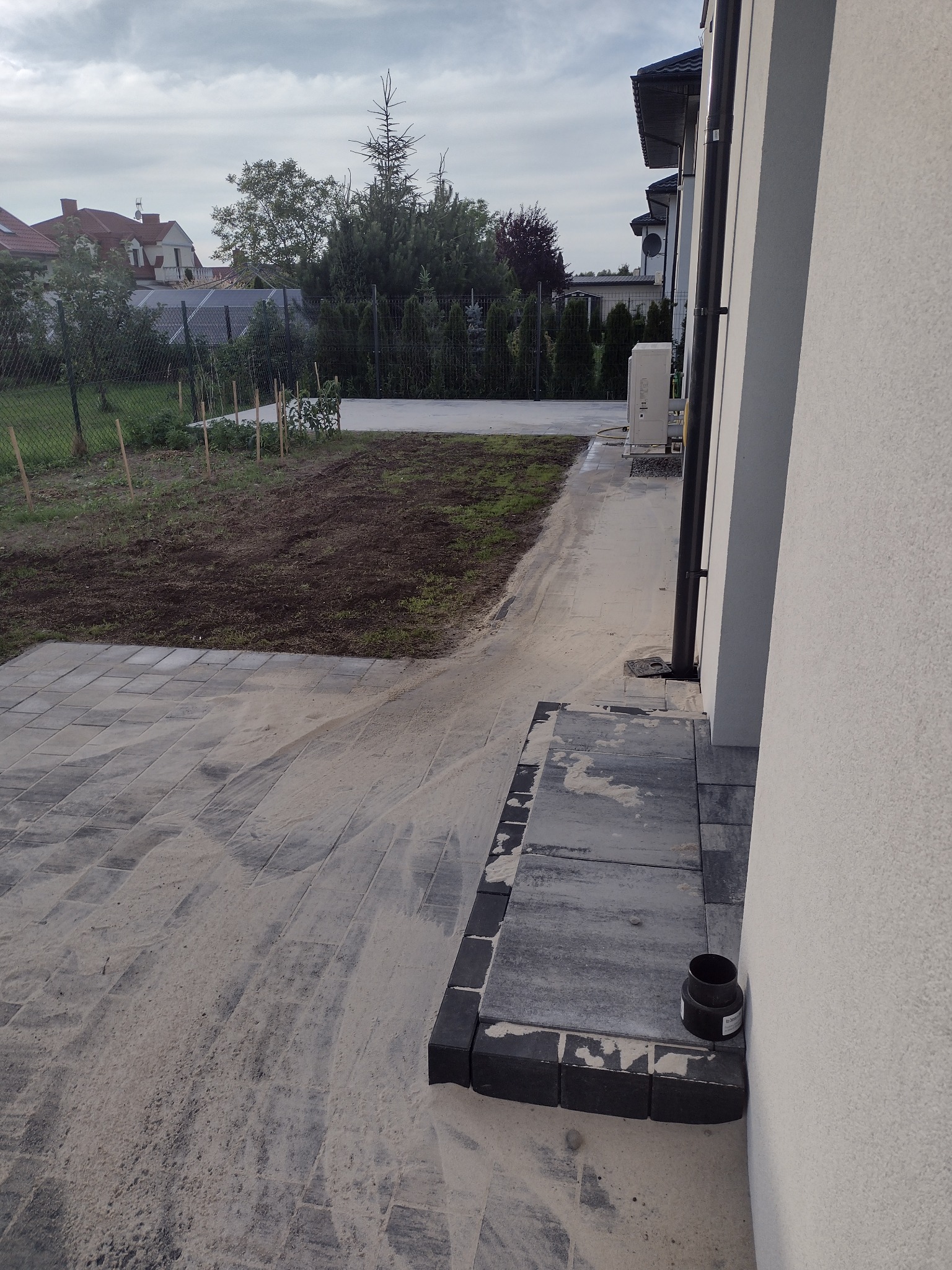 Świeżo ułożona, szara kostka brukowa przy domu, zasypana piaskiem, z widocznym fragmentem rabaty i trawnika w tle.