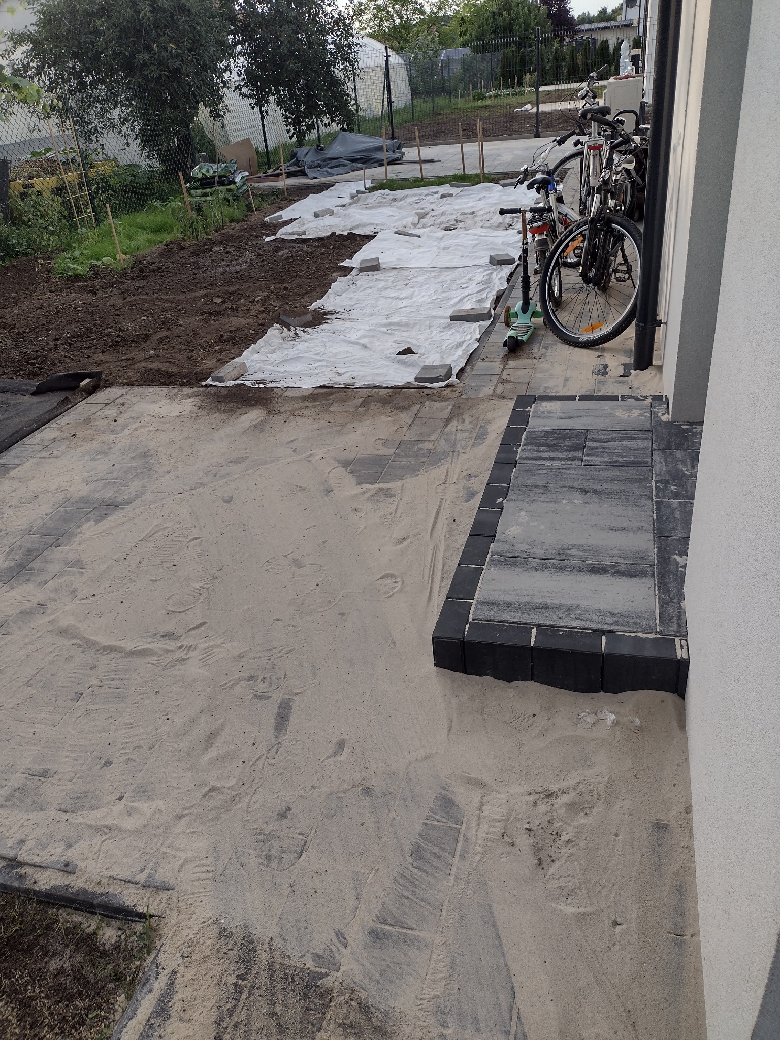 Przygotowanie podłoża pod kostkę brukową: piasek wyrównany, widoczne ślady stóp, w tle rozłożona geowłóknina i kostka, rowery i hulajnoga zaparkowane przy ścianie domu.