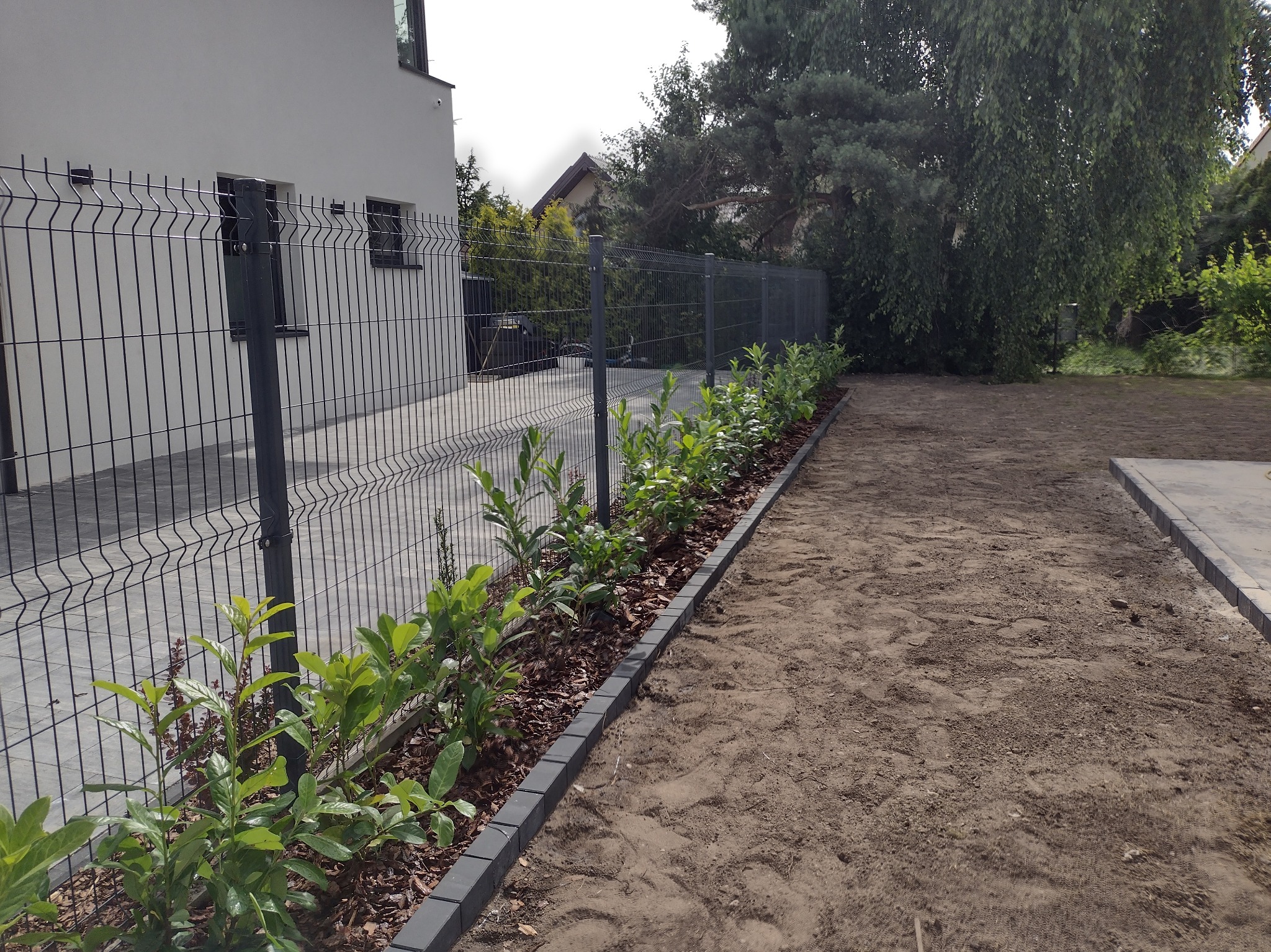 Nowoczesne ogrodzenie panelowe w kolorze grafitowym oddziela posesję od terenu z przygotowaną glebą i świeżo posadzonymi krzewami wzdłuż obrzeża z kostki.