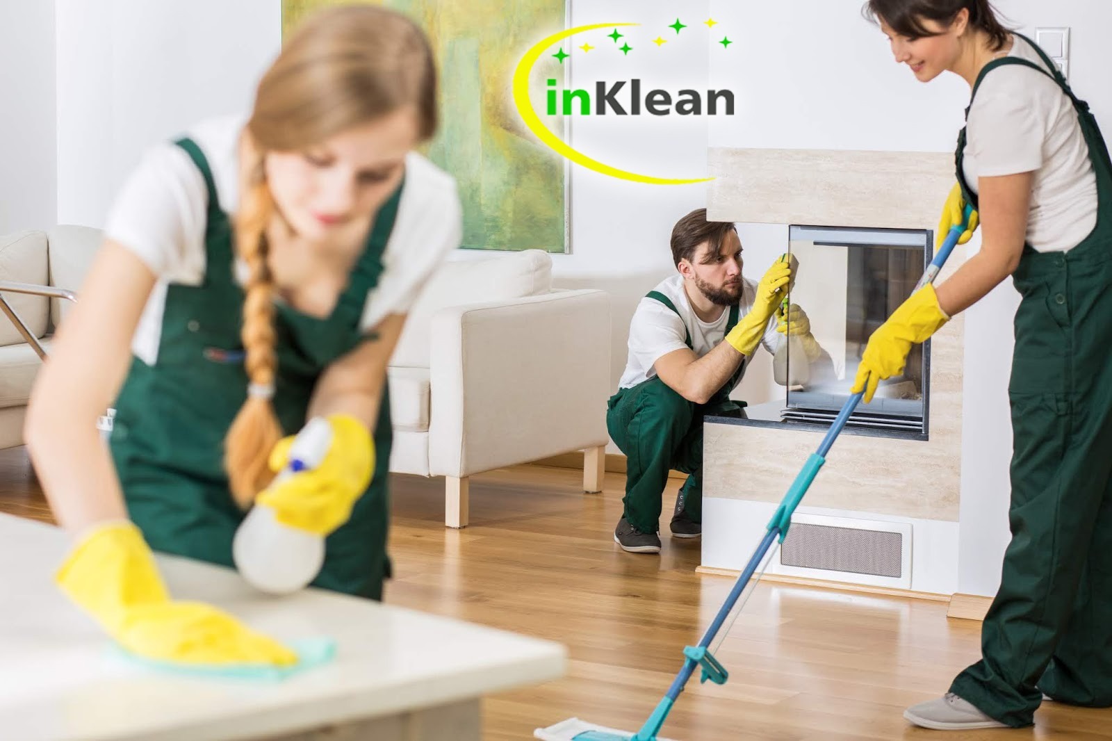 Ekipa sprzątająca w zielonych kombinezonach i żółtych rękawicach czyści kominek i stół w jasnym salonie; widoczne logo firmy InKlean.