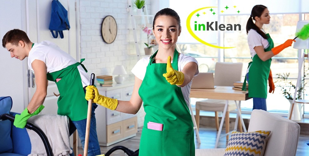 Zespół sprzątający w akcji: uśmiechnięta kobieta w centrum kadru, z uniesionym kciukiem, ubrana w zielony fartuch i żółte rękawice, w tle mężczyzna odkurzający sofę i kobieta ścierająca kurz...