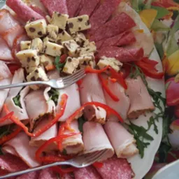 Dekoracyjnie ułożone wędliny, salami i ser z kawałkami czekolady na białym talerzu z widelcami, ozdobione czerwoną papryką i rukolą, na tle obrusu w kwiaty.