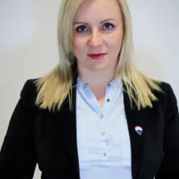 Portret kobiety w czarnej marynarce i błękitnej koszuli, z przypinką RE/MAX na klapie, uśmiechająca się do kamery na neutralnym tle.