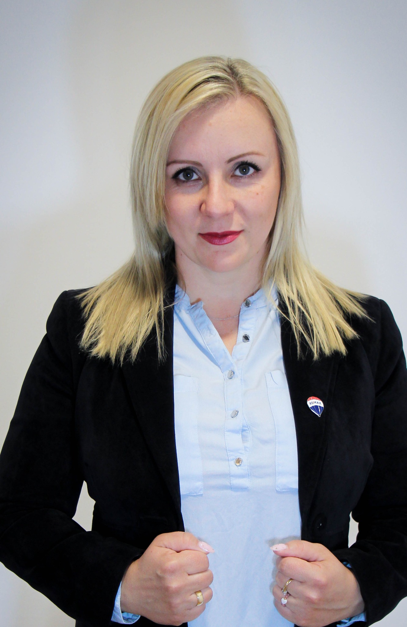 Portret kobiety w czarnej marynarce i błękitnej koszuli, z przypinką RE/MAX na klapie, uśmiechająca się do kamery na neutralnym tle.