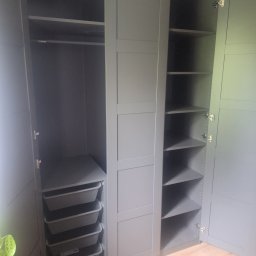 Krzysztof Roj Sprzedaż serwis naprawa - Szara szafa wnękowa z otwartymi drzwiami, ukazująca półki i wysuwane kosze. Widoczna podłoga z paneli i fragment rośliny. Ujęcie z bliska, wnętrze szafy dobrze oświetlone.