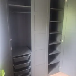 Szara szafa wnękowa z otwartymi drzwiami, ukazująca półki i wysuwane kosze. Widoczna podłoga z paneli i fragment rośliny. Ujęcie z bliska, wnętrze szafy dobrze oświetlone.