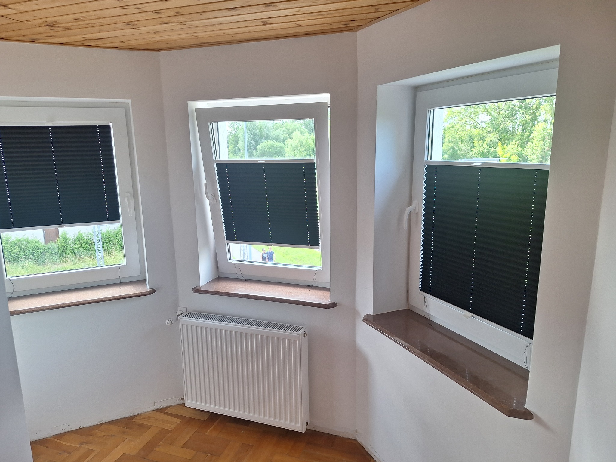Wnętrze pokoju z trzema oknami PCV z zamontowanymi czarnymi plisami okiennymi, parkiet na podłodze, drewniana boazeria na suficie i biały grzejnik pod oknem.