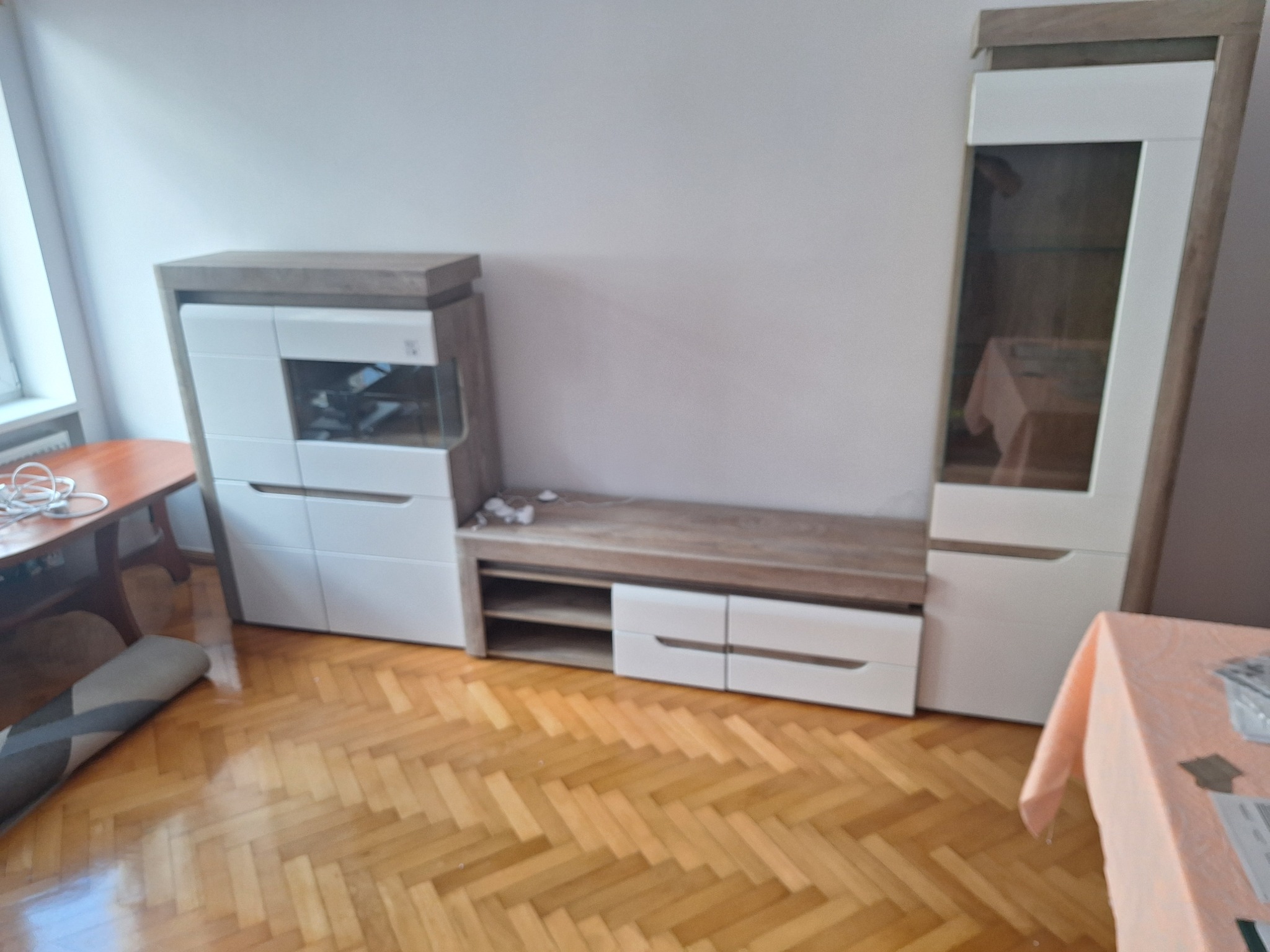 Zestaw mebli do salonu w odcieniach bieli i drewna, z parkietem ułożonym w jodełkę. Widoczna witryna z przeszkloną górną częścią i szafka RTV z szufladami. Częściowo zwinięty dywan i stół...