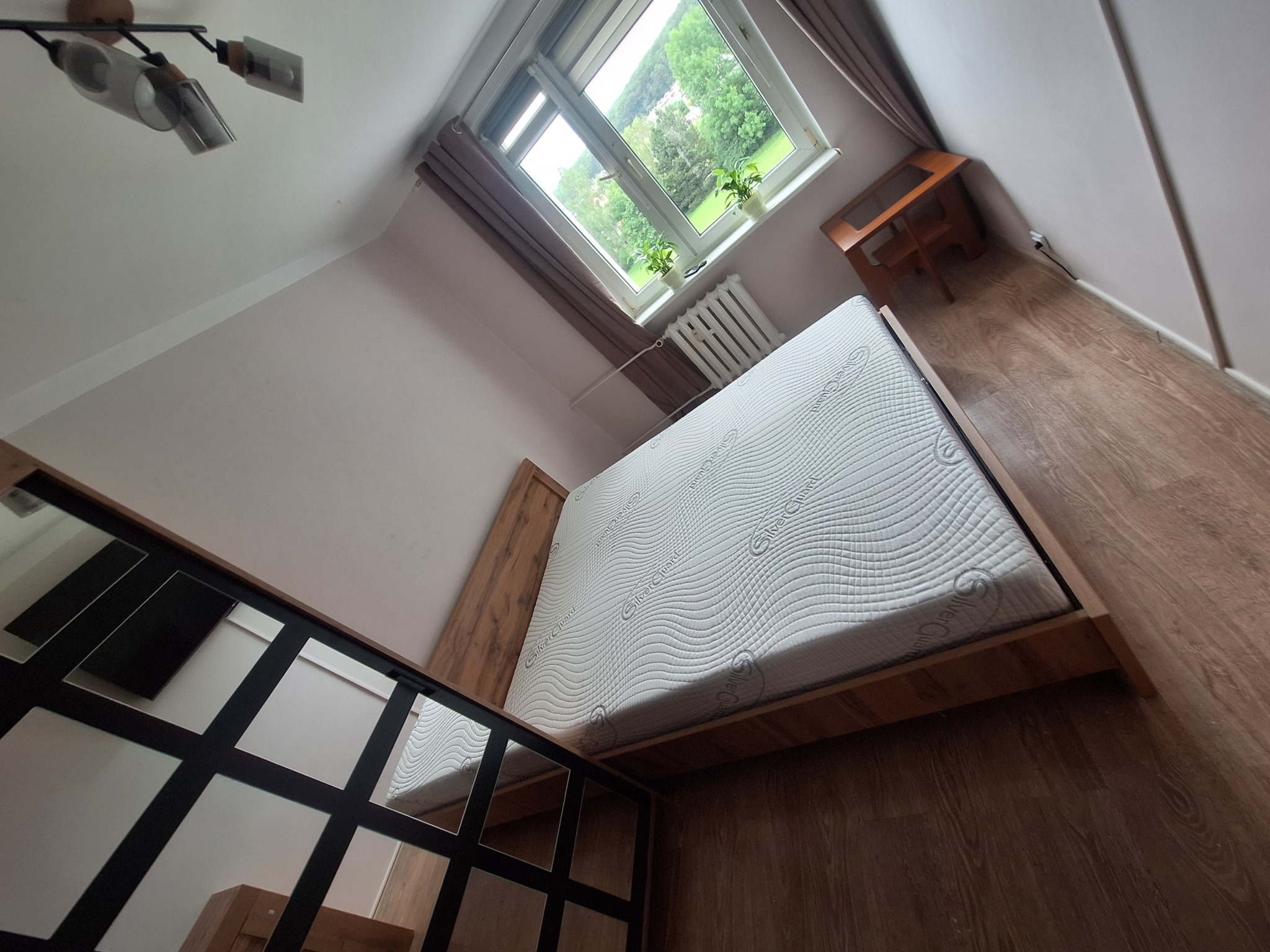 Widok na sypialnię z łóżkiem, oknem i drewnianą podłogą. Okno z roślinami w doniczkach. Nietypowa perspektywa, z góry, podkreśla przestrzeń i jasne wnętrze.