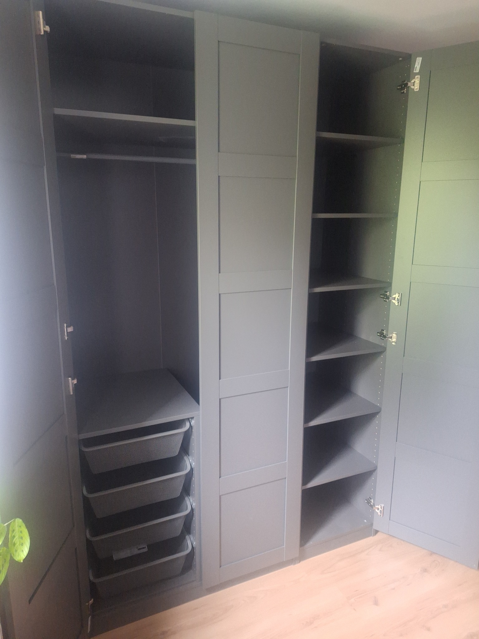 Szara szafa wnękowa z otwartymi drzwiami, ukazująca półki i wysuwane kosze. Widoczna podłoga z paneli i fragment rośliny. Ujęcie z bliska, wnętrze szafy dobrze oświetlone.