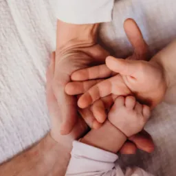 Dłonie rodziców obejmujące małą dłoń niemowlęcia na tle teksturowanej, jasnej tkaniny.