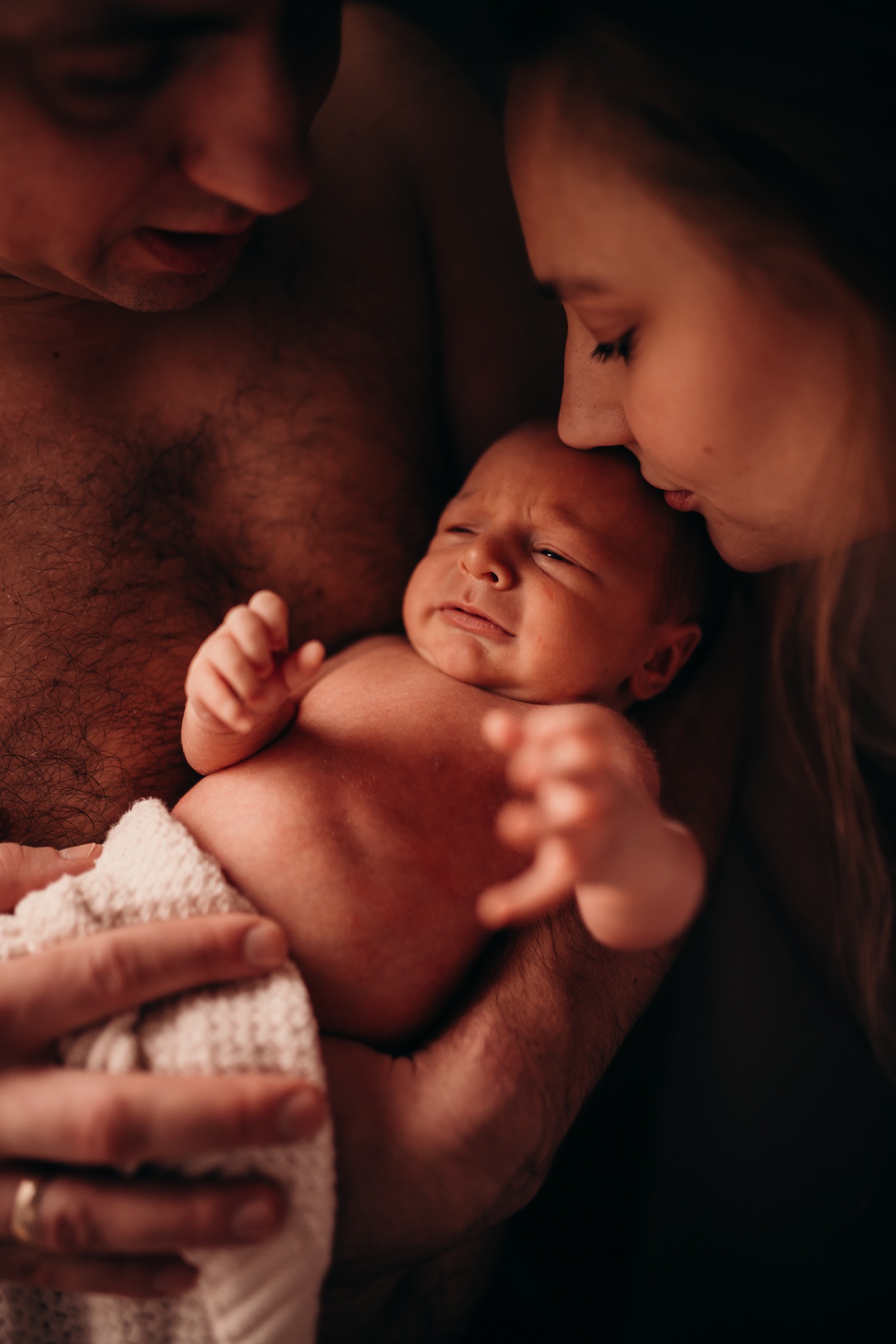 Rodzice tulą noworodka owiniętego w biały kocyk; matka całuje dziecko w czoło, ojciec trzyma w ramionach, widoczny złoty pierścionek na palcu.