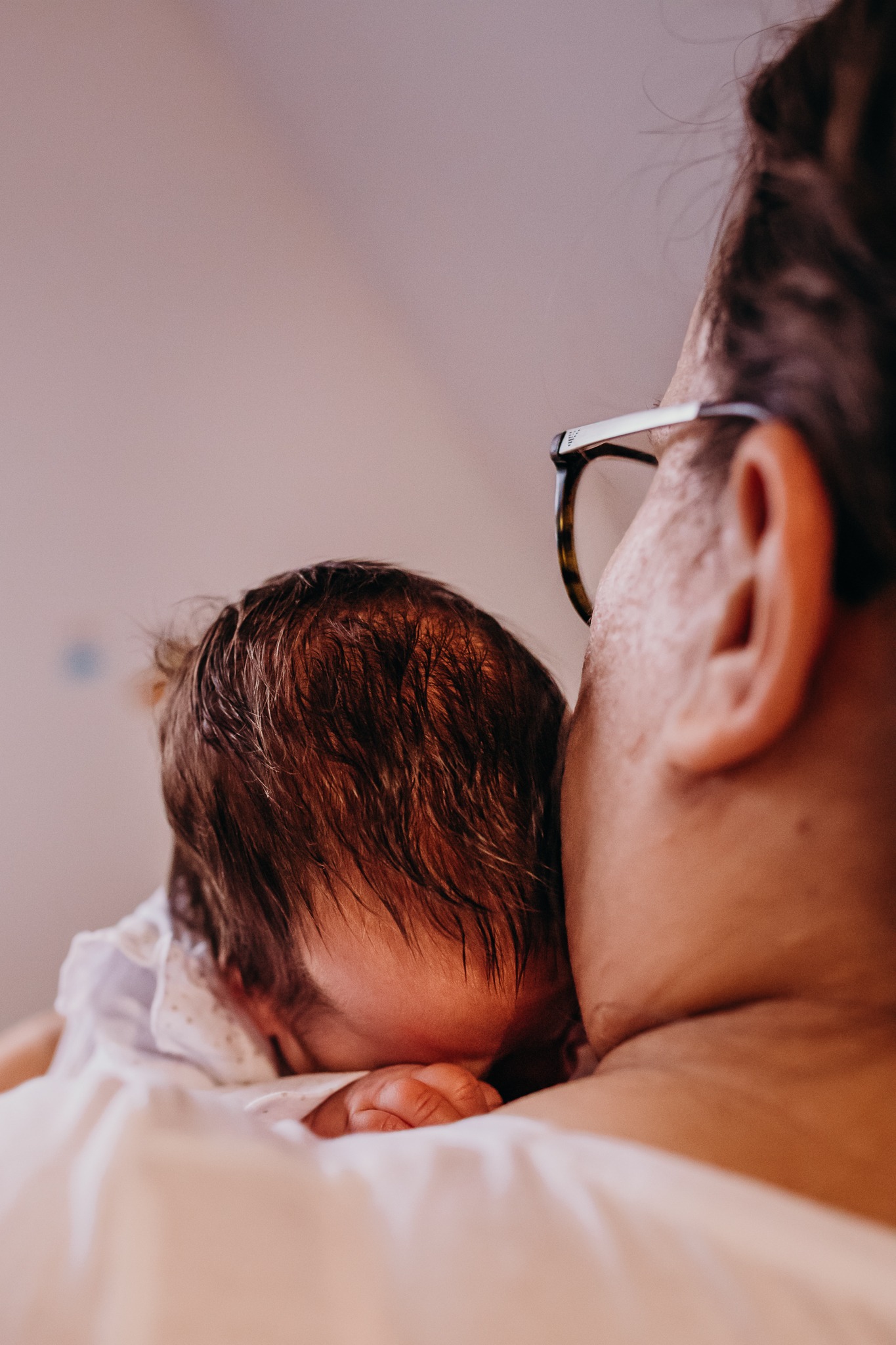 Noworodek z ciemnymi włosami śpiący na ramieniu osoby w okularach, delikatne ujęcie bliskości.