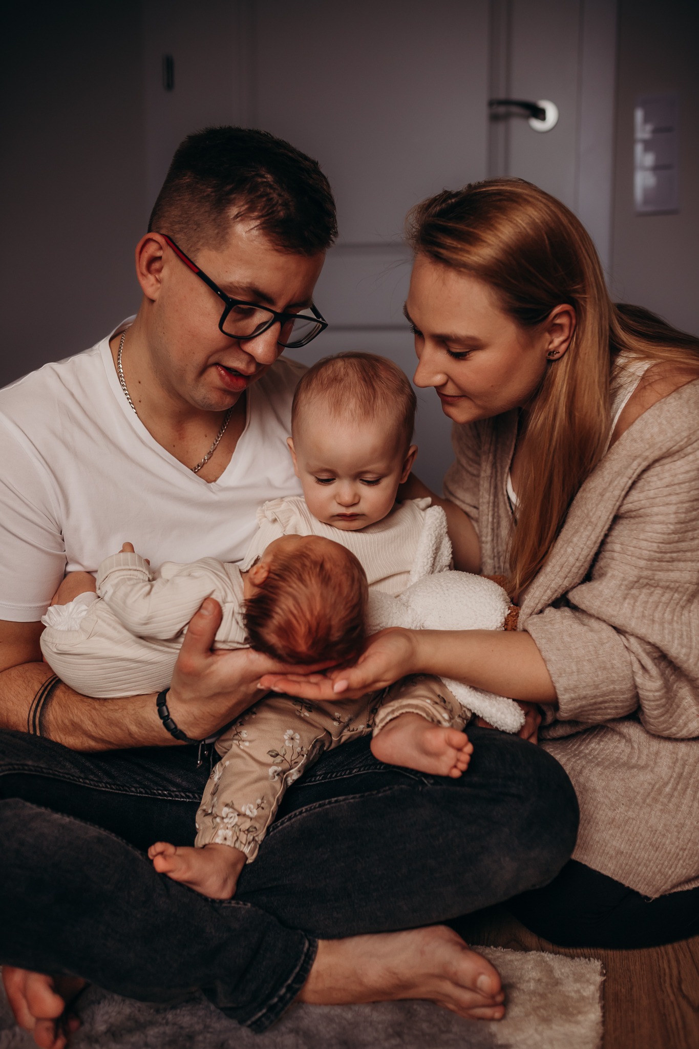 Rodzice siedzą na podłodze trzymając dwoje małych dzieci, jedno w ramionach ojca, drugie oparte o matkę, z bliska widoczne stopy dziecka na kolanach ojca.