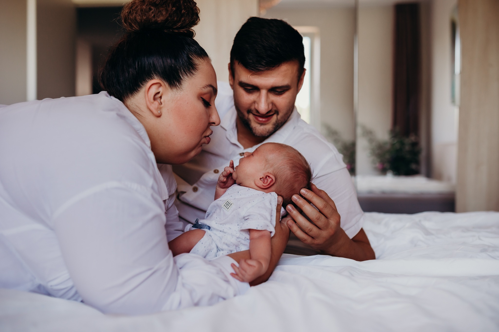 Mama z tatą czule spoglądają na noworodka w Piasecznie, leżąc na białej pościeli, delikatne światło podkreśla bliskość rodziny.