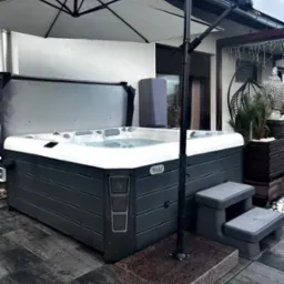 Zewnętrzne jacuzzi z otwartą pokrywą, umieszczone na szarym tarasie z kamiennej kostki, osłonięte białym parasolem na tle białego domu z ciemną stolarką okienną i dekoracyjną figurką Buddy.