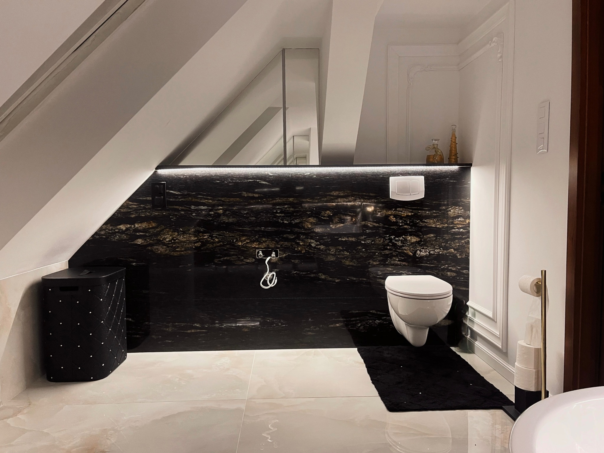 Elegancka łazienka na poddaszu z czarną ścianą z kamienia naturalnego z złotymi żyłkami, wisząca toaleta i marmurowa podłoga w beżowych odcieniach.