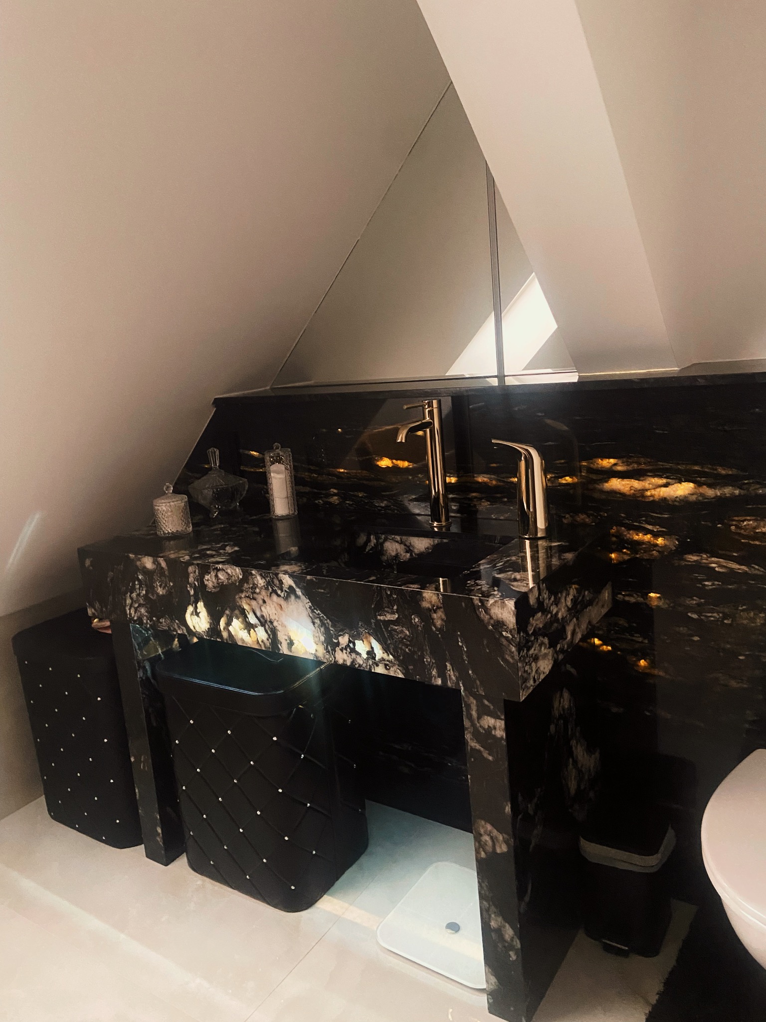 Czarny, marmurkowy blat łazienkowy z dwiema złotymi bateriami umywalkowymi, dopasowany do ściany, z czarnymi akcesoriami łazienkowymi w pomieszczeniu ze skośnym sufitem.