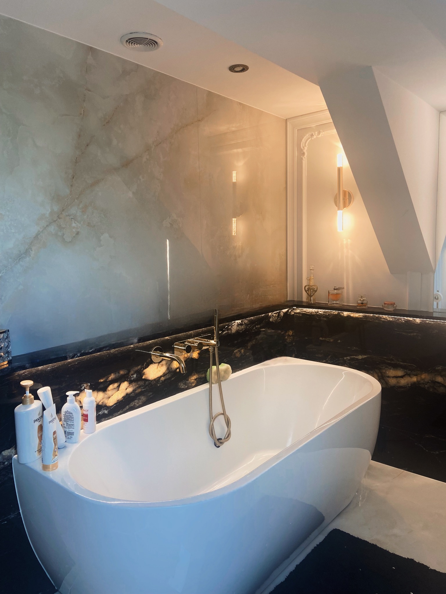 Nowoczesna łazienka z wolnostojącą białą wanną, czarnym blatem z wyraźnym żyłkowaniem i beżową okładziną ściany imitującą marmur, oświetlona dekoracyjnymi lampami.