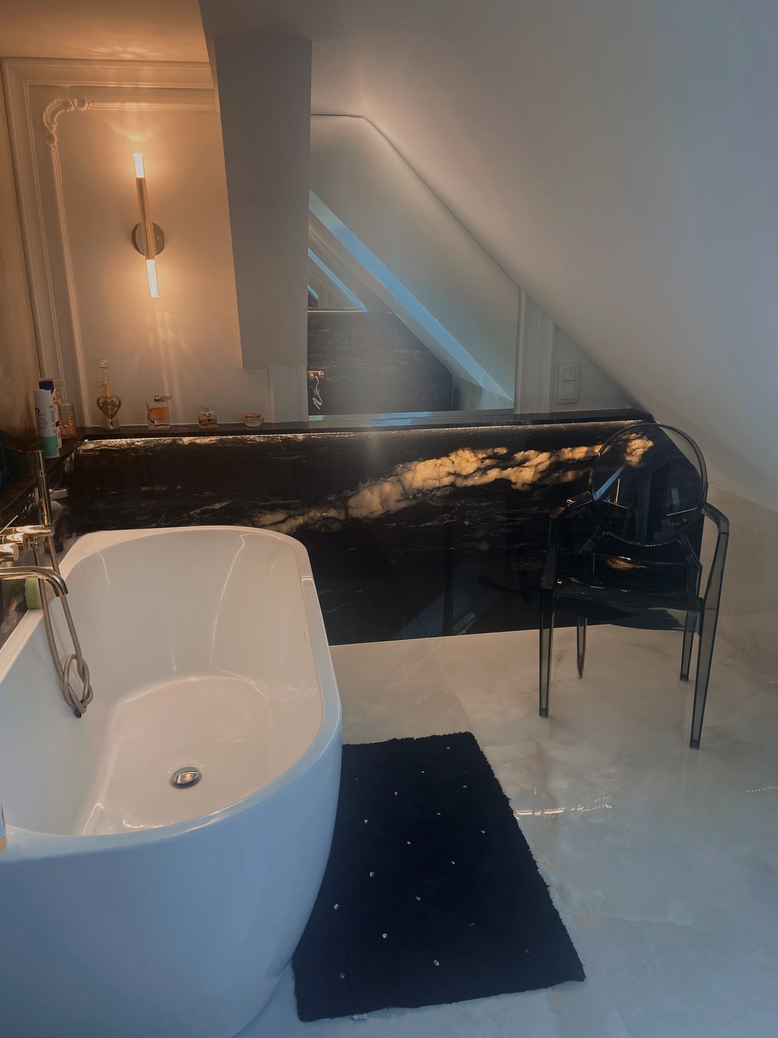 Nowoczesna łazienka na poddaszu z czarnym blatem o marmurkowym wzorze, białą wanną wolnostojącą i transparentnym krzesłem.