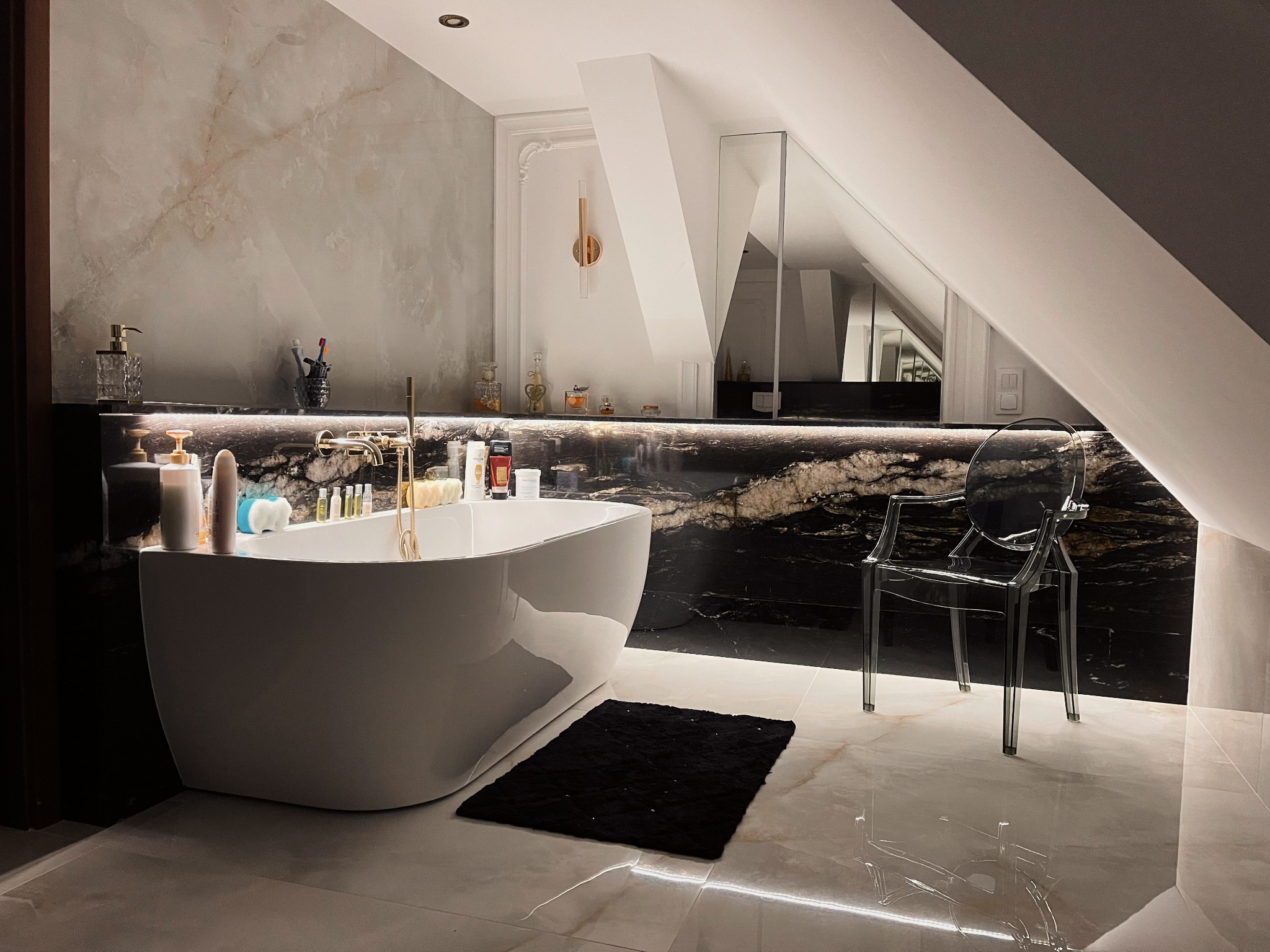 Nowoczesna łazienka na poddaszu z marmurową ścianą, białym wolnostojącym zlewem, złotą armaturą i transparentnym krzesłem.