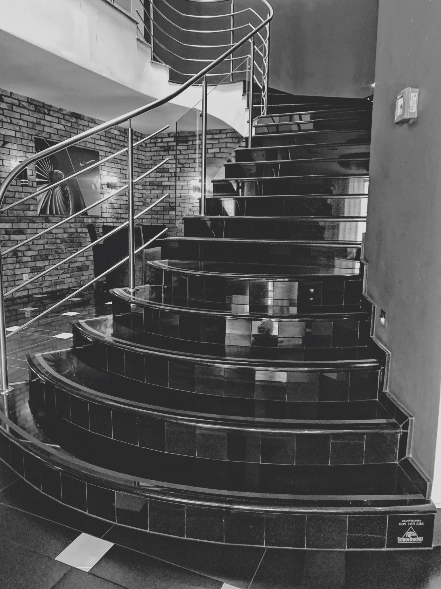 Spiralne schody z czarnego kamienia z metalową balustradą, widoczne logo firmy na dole schodów, ściana z cegły w tle, czarno-białe zdjęcie.