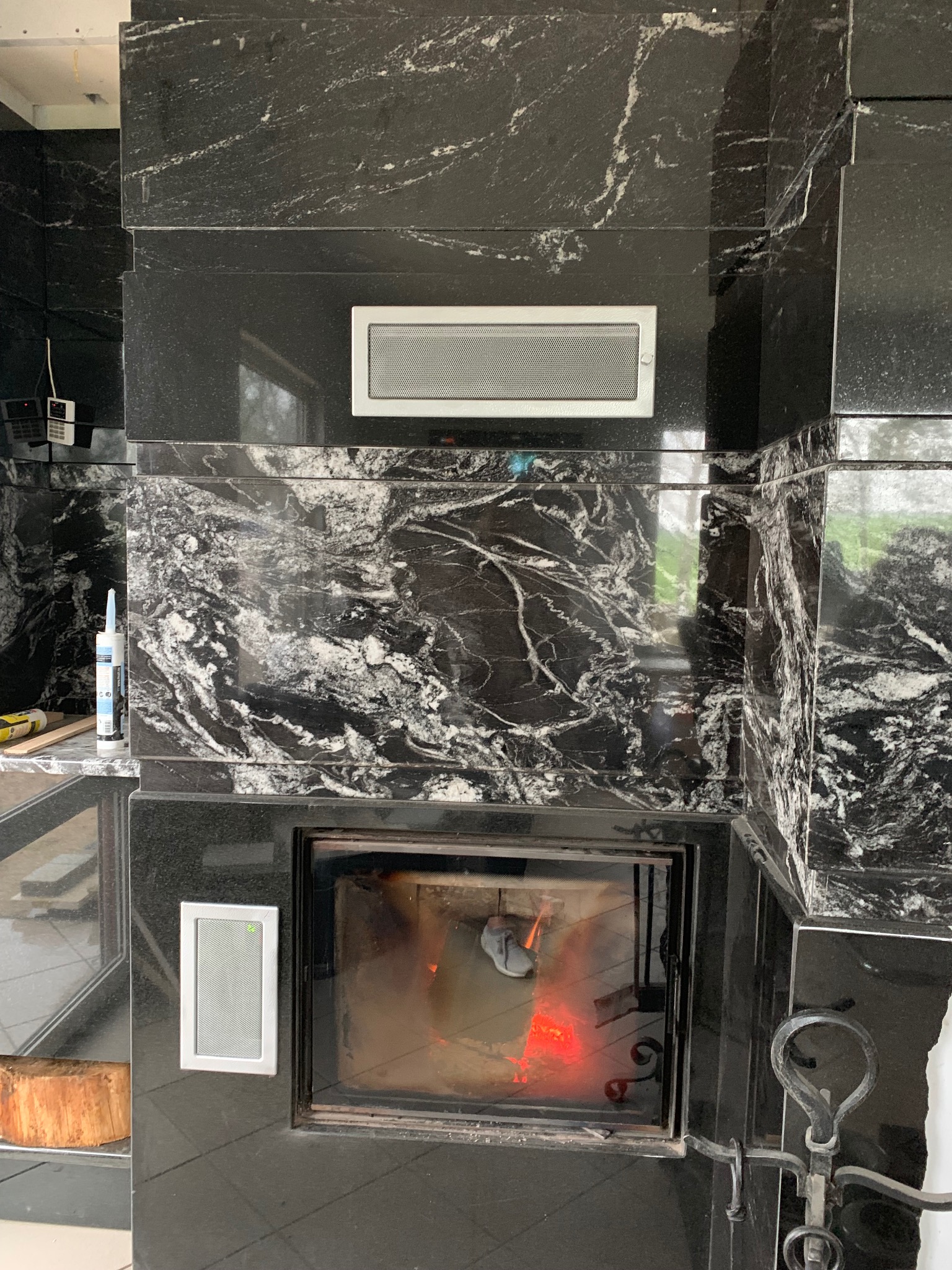 Obudowa kominka wykończona czarnym kamieniem z białymi żyłami, widoczne elementy montażowe i akcesoria kominkowe, płomienie wewnątrz paleniska.