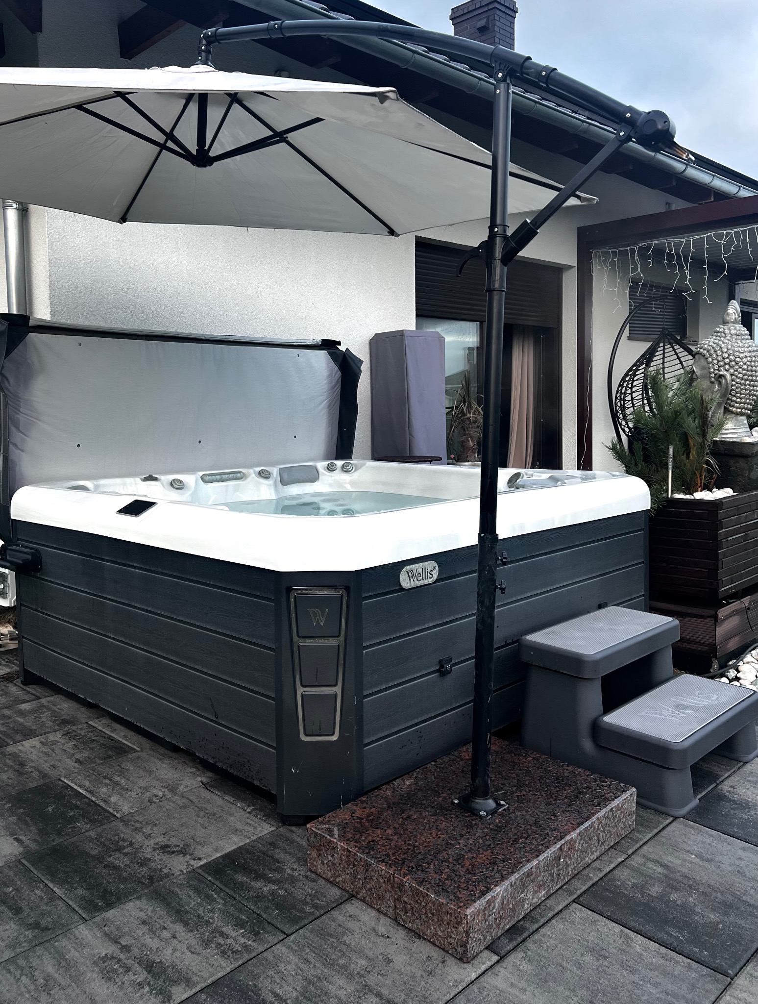 Zewnętrzne jacuzzi z otwartą pokrywą, umieszczone na szarym tarasie z kamiennej kostki, osłonięte białym parasolem na tle białego domu z ciemną stolarką okienną i dekoracyjną figurką Buddy.