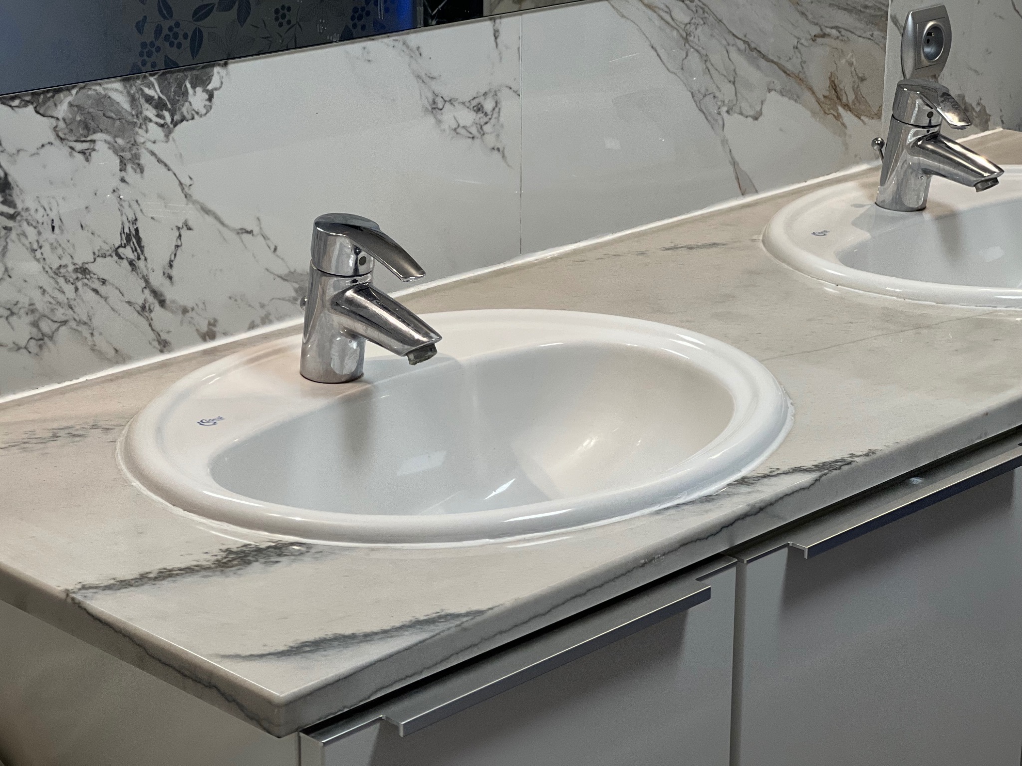 Dwie białe umywalki nablatowe z chromowanymi bateriami, osadzone w blacie z konglomeratu o wzorze marmuru, zamontowane na szarych szafkach łazienkowych.