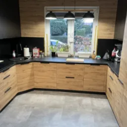 Nowoczesna kuchnia w kształcie litery U z frontami szafek w kolorze jasnego drewna i czarnymi blatami, oświetlona trzema lampami wiszącymi nad oknem z widokiem na ogród.