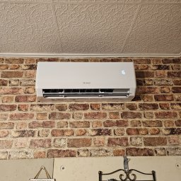 Sun House 🌞 - Biały klimatyzator marki Gree zamontowany na ścianie z cegieł, wyświetlający temperaturę 16 stopni Celsjusza, widoczny fragment dekoracyjnego sufitu kasetonowego.