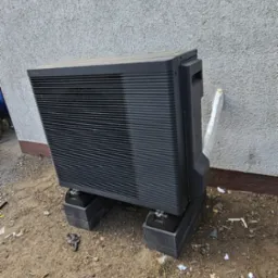 Zewnętrzna jednostka klimatyzacyjna marki Panasonic zamontowana na betonowych podstawach przy szarej ścianie budynku, z widoczną izolacją rury.