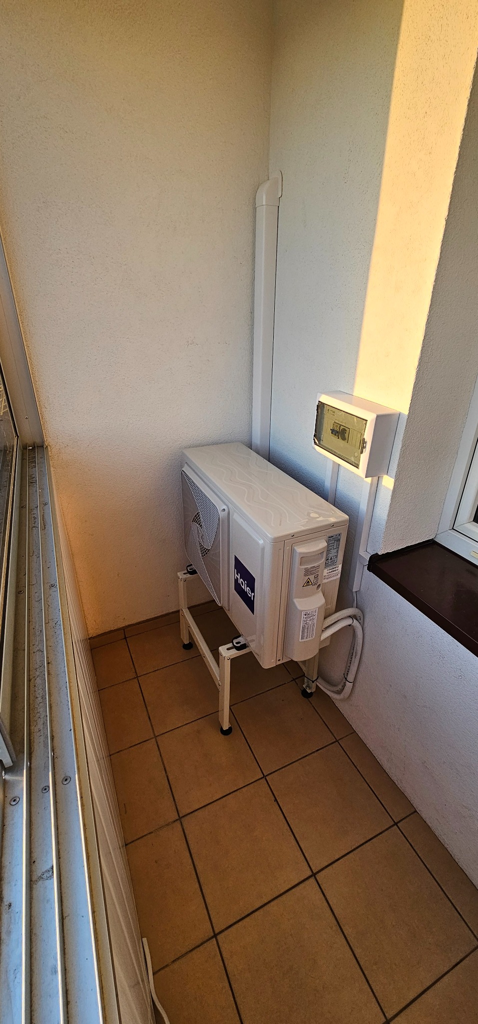 Zewnętrzna jednostka klimatyzatora marki Haier, zamontowana na metalowym stelażu na balkonie z brązową terakotą, widoczne rury i skrzynka elektryczna.