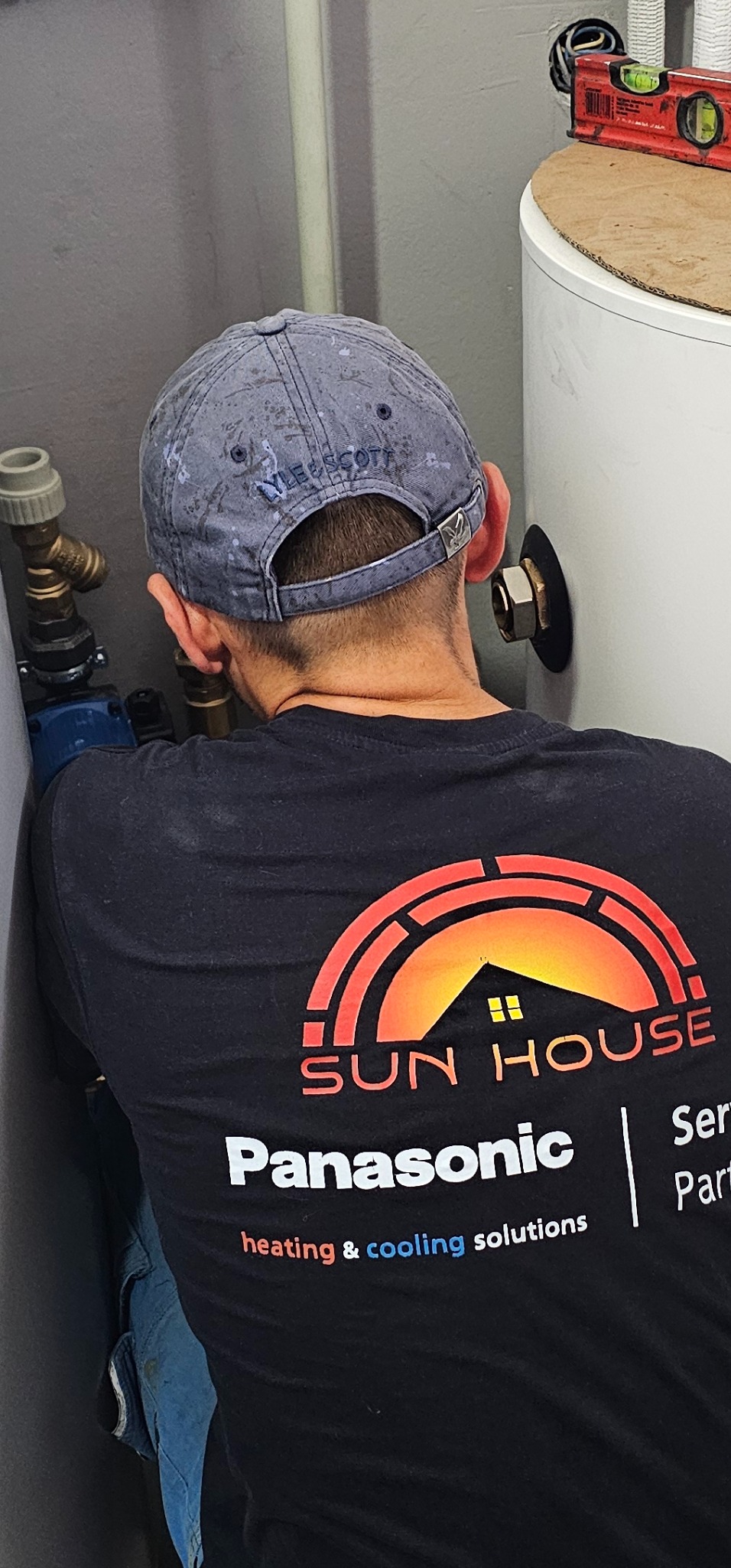 Instalator w Toruniu w trakcie podłączania instalacji do zasobnika ciepłej wody, widoczny poziomica na górze zasobnika, instalator ma na sobie koszulkę firmy Sun House Panasonic heating & cooling...